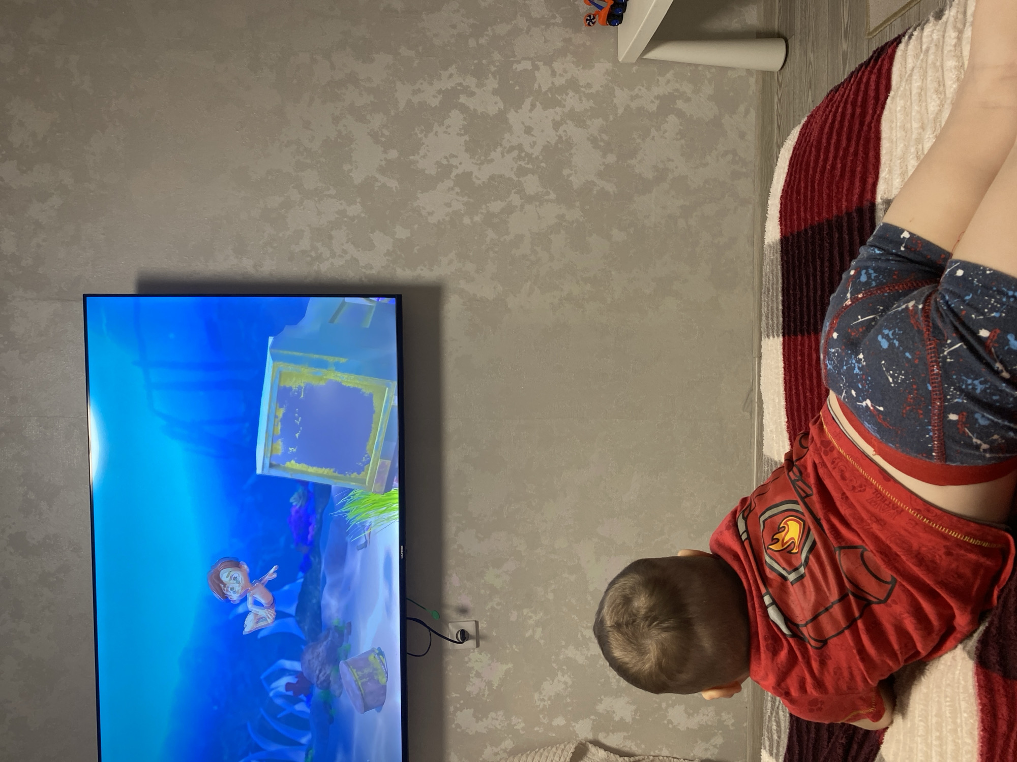 An overhead shot captures a young child lying face down on a red and white striped blanket, intently watching a large flat-screen television. The child is seen from behind, revealing the back of their head with short brown hair, a red t-shirt adorned with a shield-like emblem featuring a fire graphic, and blue shorts with a white, red, and blue speckled pattern.
The television screen displays a vibrant animated scene, appearing to be from a video game or cartoon. It features a cartoon character resembling a young boy with brown hair and light skin, depicted bare-chested in a vivid blue underwater environment. Various underwater structures and plant life are also visible on the screen. The brand "SAMSUNG" is faintly visible on the bottom bezel of the television.
The wall behind the television and the child is covered in a light gray or beige wallpaper with a distinct, mottled texture. A small white electrical outlet with black cables connected to the TV is located on this wall. A portion of a white shelf or furniture piece is visible in the upper right corner of the frame. The overall setting is an indoor, comfortable space, likely a living room or bedroom, where the child is engrossed in screen time.