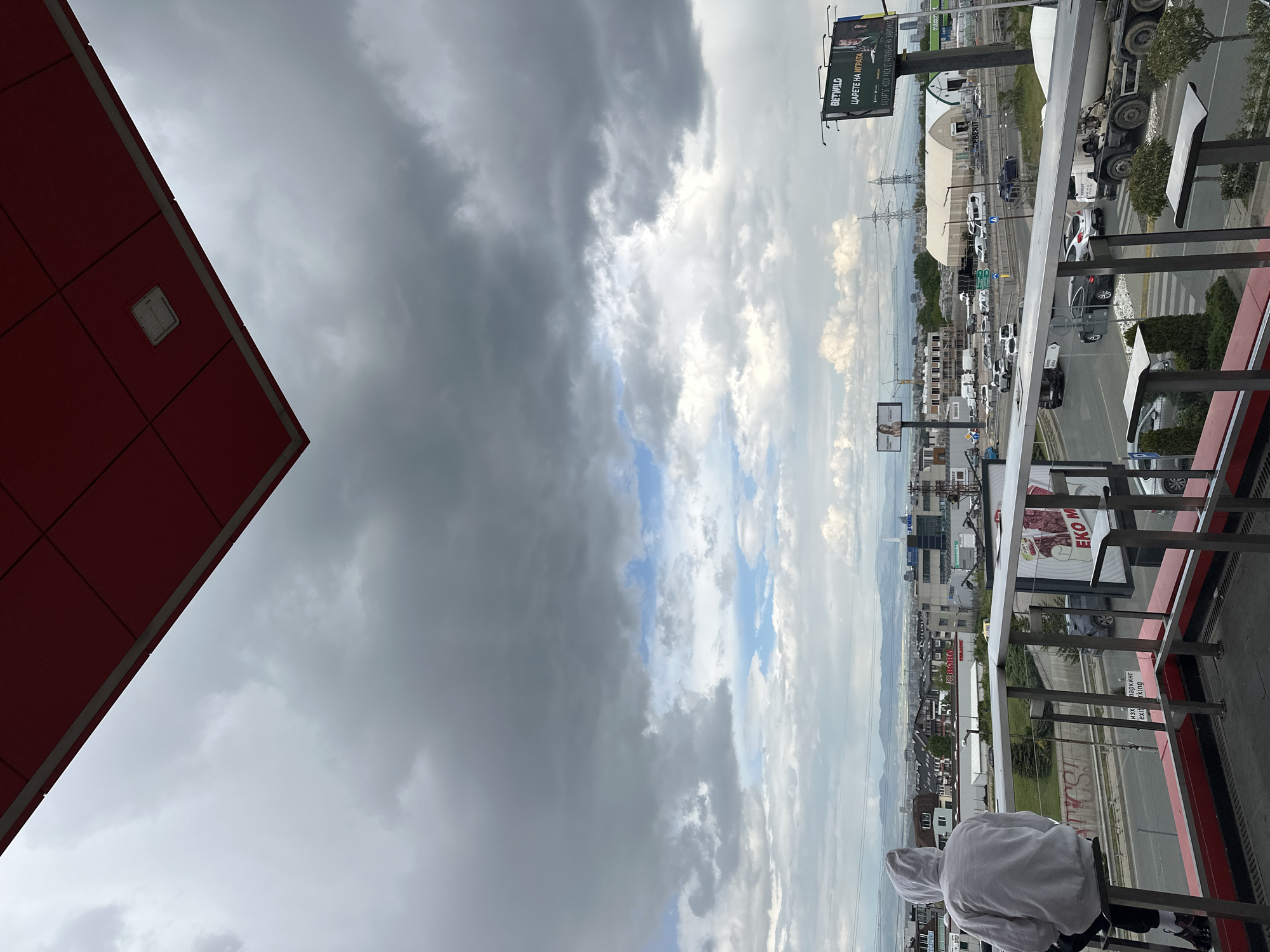 The image shows an elevated outdoor view, likely from a balcony, overlooking a cityscape, probably in Sofia, Bulgaria. To the left, a portion of a red building is visible. The foreground includes balcony railings and a person in a light-colored hooded sweatshirt, facing away from the camera. The midground features a busy road with vehicular traffic, including cars and a cement truck. There are also billboards with advertising present, and some buildings, one of which says "Liberty." Beyond the road, the city extends into the background, with residential buildings interspersed with some green spaces. The sky is filled with dark, ominous storm clouds mixed with patches of lighter clouds and some blue sky. Electrical power lines stretch across the cityscape.
