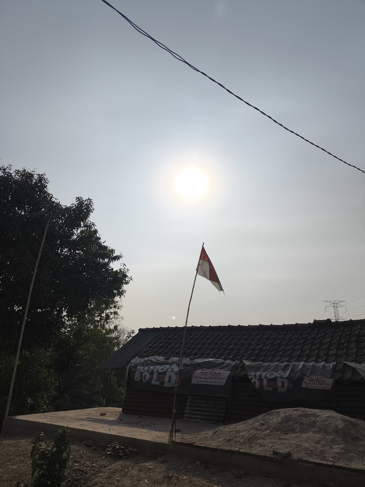 The image depicts an outdoor scene in Cilegon, Indonesia, on a clear day with the sun prominently visible in the sky. In the foreground, a pile of sand is situated next to a structure with a tiled roof, possibly under construction or storage. The structure is partially covered with plastic sheeting that has text and price markings, including "Rp 25.000 an". To the left of the structure, a tree with dense foliage stands tall. A thin pole with the Indonesian flag, red and white, is planted in front of the structure, slightly to the right of the tree. In the background, a power transmission tower can be seen in the distance. Overhead, two wires stretch across the sky, suggesting the presence of power lines. The overall impression is one of a semi-urban or rural setting in Cilegon, Indonesia, captured during daylight hours.