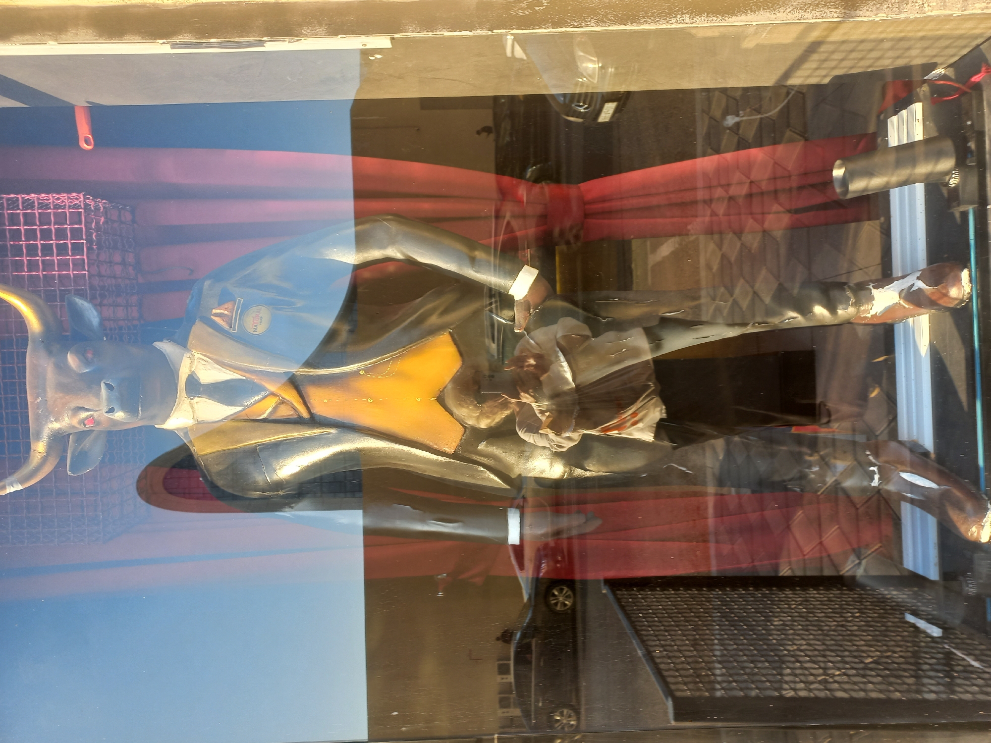 The image shows a display window with a mannequin of a bull wearing a suit. The bull has red eyes and gold horns. It is dressed in a black suit, a yellow vest, a white shirt, and a tie. A badge with the word "NATURAL" is visible on its lapel. Behind the bull, red curtains are drawn.
Reflected in the glass, a woman holding a phone is visible, appearing to take a photograph of the display. Also reflected are cars parked on a street, a fence, and a building facade. The scene is set on a street in Baku, Azerbaijan, with the golden hour light illuminating the exterior. The time of day appears to be late afternoon or early evening, given the warm lighting.