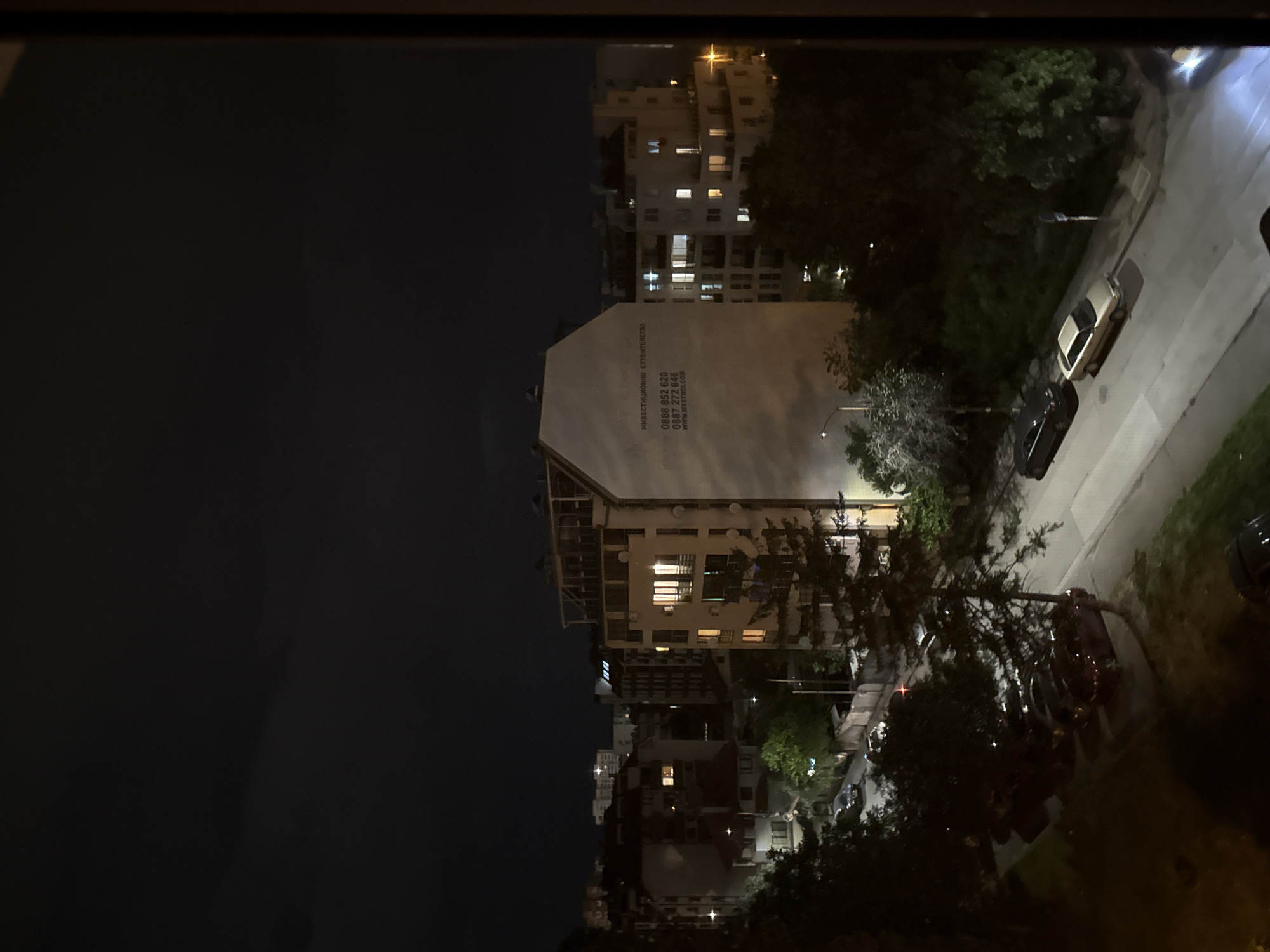 The image depicts a night scene in Sofia, Bulgaria, featuring buildings and a street. A building with the inscription "ИНВЕСТИЦИОННО СТРОИТЕЛСТВО" and phone numbers is prominently visible. Several cars are parked along the street. The image includes trees and streetlights. A dark sky is above the buildings.