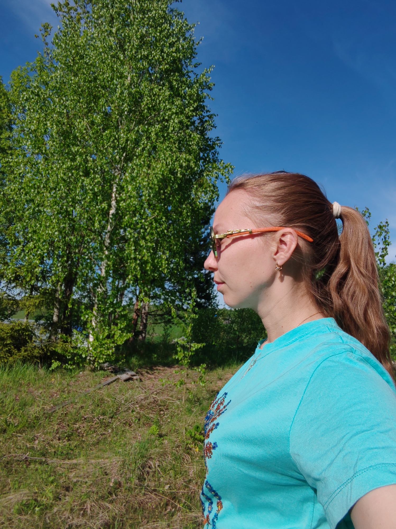 A woman is seen in profile, wearing glasses and a blue t-shirt, standing in an outdoor area. There are trees and grass behind her. The location is Чухлэм, Russia.