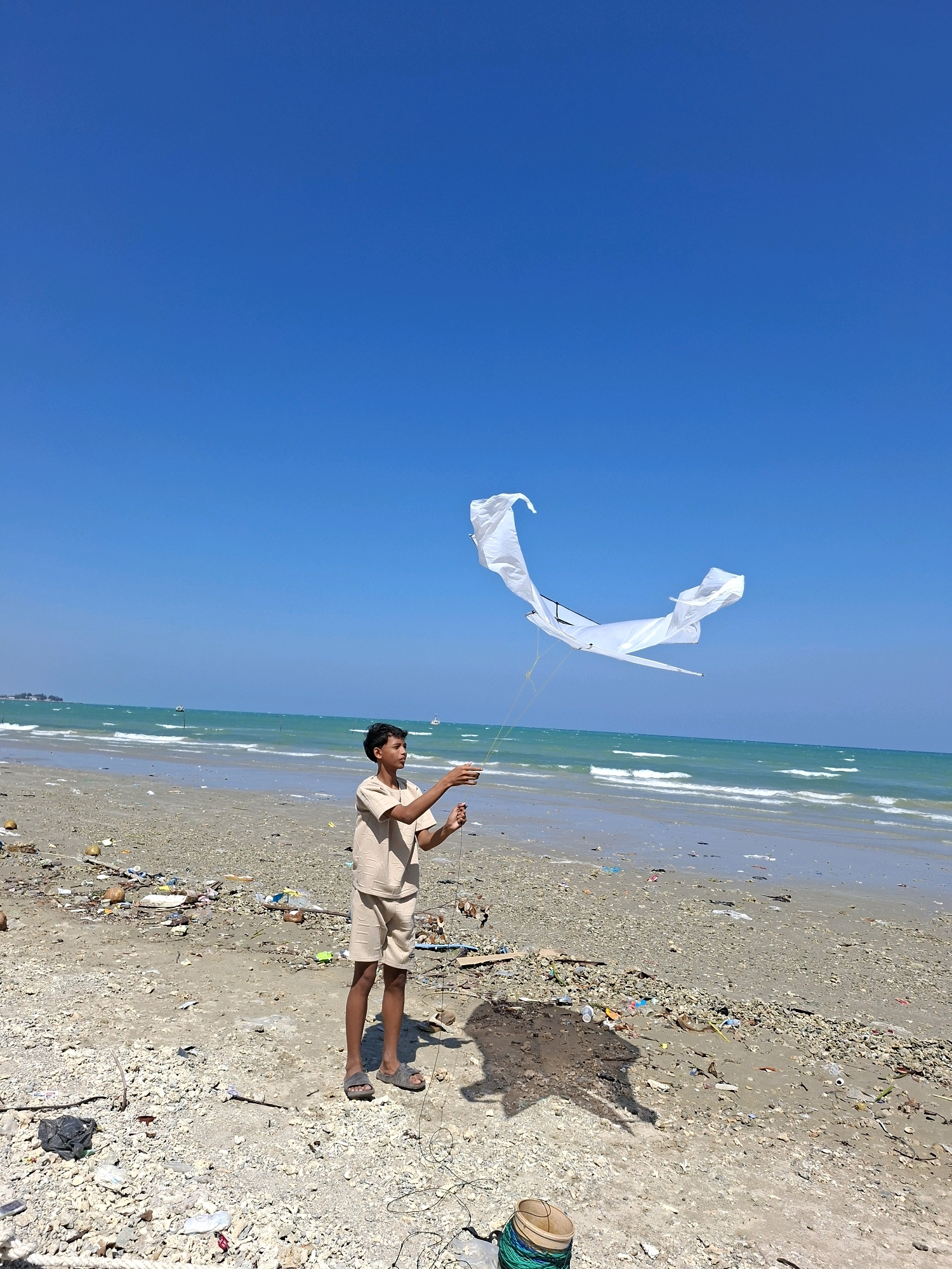 A young boy stands on a sandy beach, holding the string of a white kite in the air. The kite is a large, flowing structure with multiple fabric panels that catch the wind. The beach is littered with debris, including plastic bottles, bags, and other trash, which extends to the edge of the water. The ocean has gentle waves, and the sky is clear and blue. In the distance, there are faint outlines of boats. The boy is wearing a light-colored short-sleeved shirt and shorts. He is wearing sandals. The scene appears to be the city of Camplong, Indonesia.