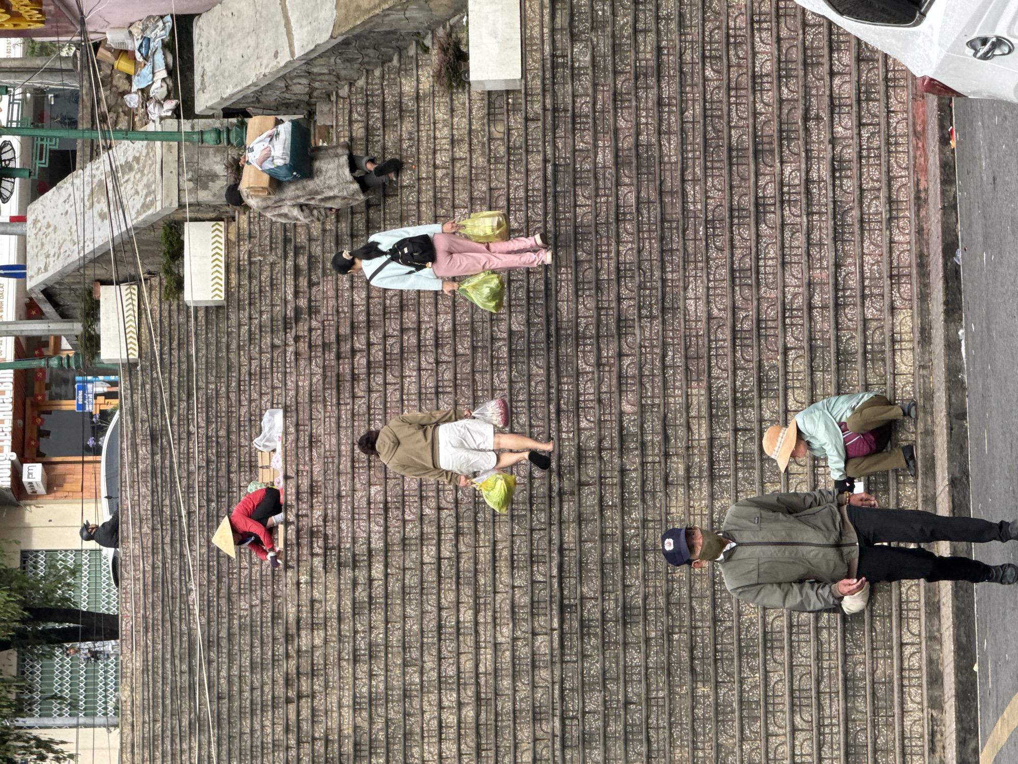 An overhead view captures a bustling urban scene on a wide, tiled outdoor staircase or steep pedestrian street in Da Lat, Vietnam. The steps are paved with dark, patterned tiles.
Multiple people are present, engaged in daily activities. In the upper left, a person is resting or sleeping, wrapped in a blanket, next to cardboard boxes and other items on a landing. Below them, a woman wearing a light blue top, pink pants, and a black backpack walks down the steps, holding two yellow plastic bags. Further down, a man in a khaki jacket and white shorts also walks with a yellow plastic bag and another bag containing red produce, possibly flowers.
Towards the bottom left, a person wearing a red jacket and a traditional conical straw hat (nón lá) is kneeling beside a wooden crate, seemingly preparing or selling goods. In the lower right, an older man in a green jacket, black pants, baseball cap, and face mask stands on the steps, holding a light-colored cloth. Next to him, another individual in a light green shirt and a straw hat is bent over, attending to something on the ground. Buildings with visible shopfronts and metal security grilles line the left side of the street, while a road with a partially visible white car borders the right.
The scene depicts everyday life in a public space, suggesting people are commuting, shopping, or working. The lighting indicates it is daytime, likely overcast as shadows are soft. White concrete blocks with yellow chevron patterns are visible on some steps. Visible text on signs includes fragmented Vietnamese, such as "...CA BINH DA LAT..." and a number "...3.623.157...", along with "pm" on another sign.