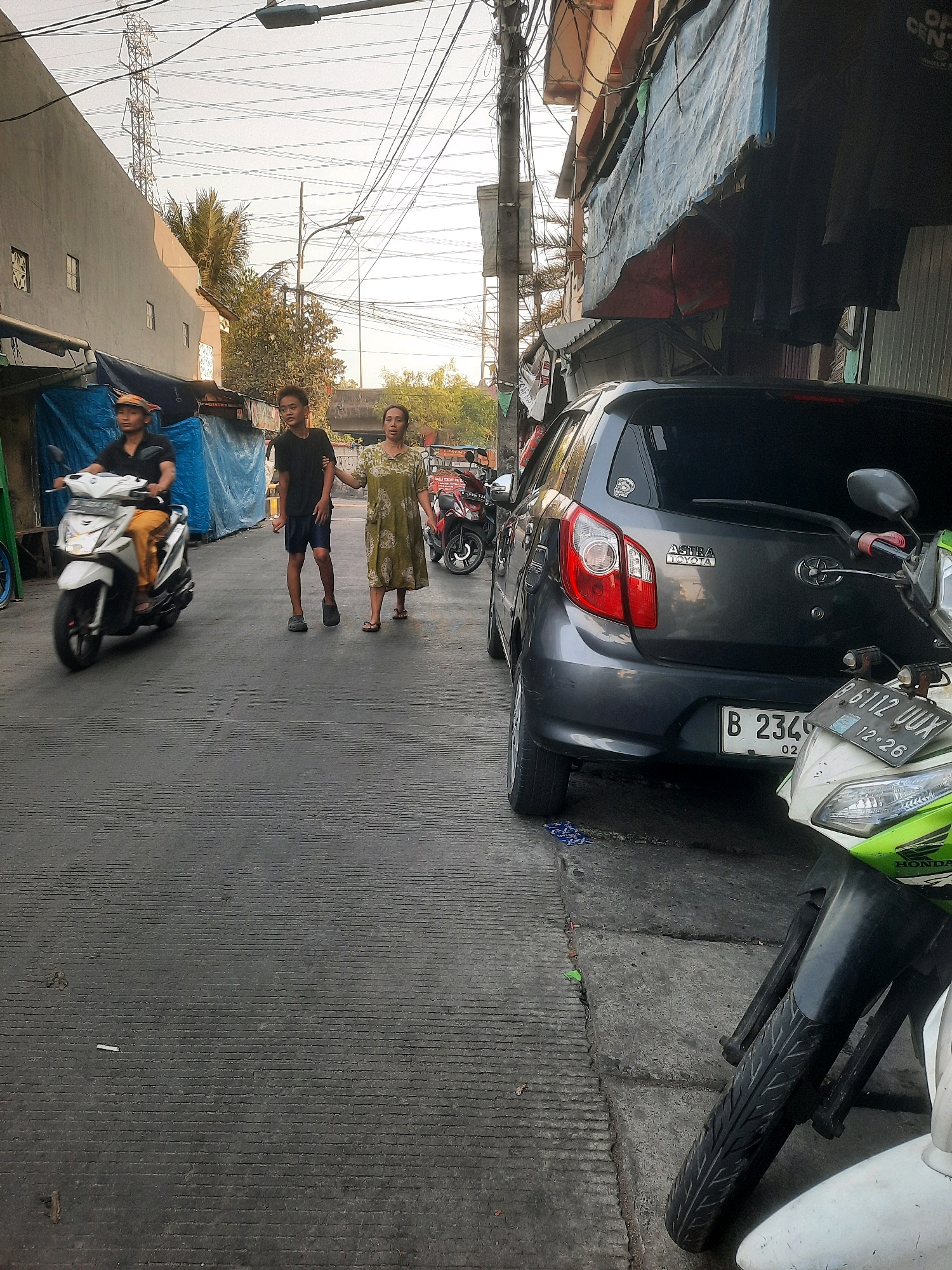 In a narrow urban street in North Jakarta, Indonesia, a person rides a white scooter from left to right. Further down the street, two individuals, a young male and an adult female, walk hand-in-hand from right to left, with the female appearing to guide the male. A dark gray Toyota car is parked on the right side of the street, with its rear bumper and taillights visible. In the foreground on the right, the front of a white and green motorcycle is parked, partially obscuring the car. The street is paved with dark asphalt, showing some texture. Buildings line both sides of the street, with tarps and hanging items visible on the right. Overhead, a dense network of power lines crisscrosses the sky. The lighting suggests it is late afternoon or early evening, casting a soft light on the scene.