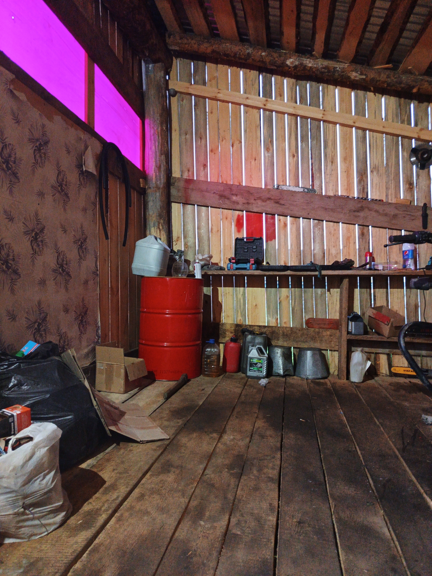 The image depicts the interior of a rustic wooden structure, likely a workshop or storage area, in the village of Islambaevo, Russia. The scene is illuminated by a bright pink light filtering through a window or opening at the top left, casting a vibrant hue on the wall and ceiling. The walls are constructed of vertical wooden planks, and the ceiling appears to be made of similar material, with exposed beams.
On the left side of the image, a patterned wallpaper covers a section of the wall. Near the floor, a large black garbage bag and a white-bagged item are present. A red barrel stands prominently in the center of the frame, topped with a light-colored plastic container. To the right of the barrel, various items are stored on wooden shelves. These include containers of oil or other liquids, buckets, and what appears to be a toolbox. A red bucket and a clear plastic bottle are also visible on the floor. In the background, a chainsaw is partially visible. The floor is made of worn wooden planks. The overall impression is one of a utilitarian space that is somewhat cluttered with tools and supplies. There are no people visible in the image.