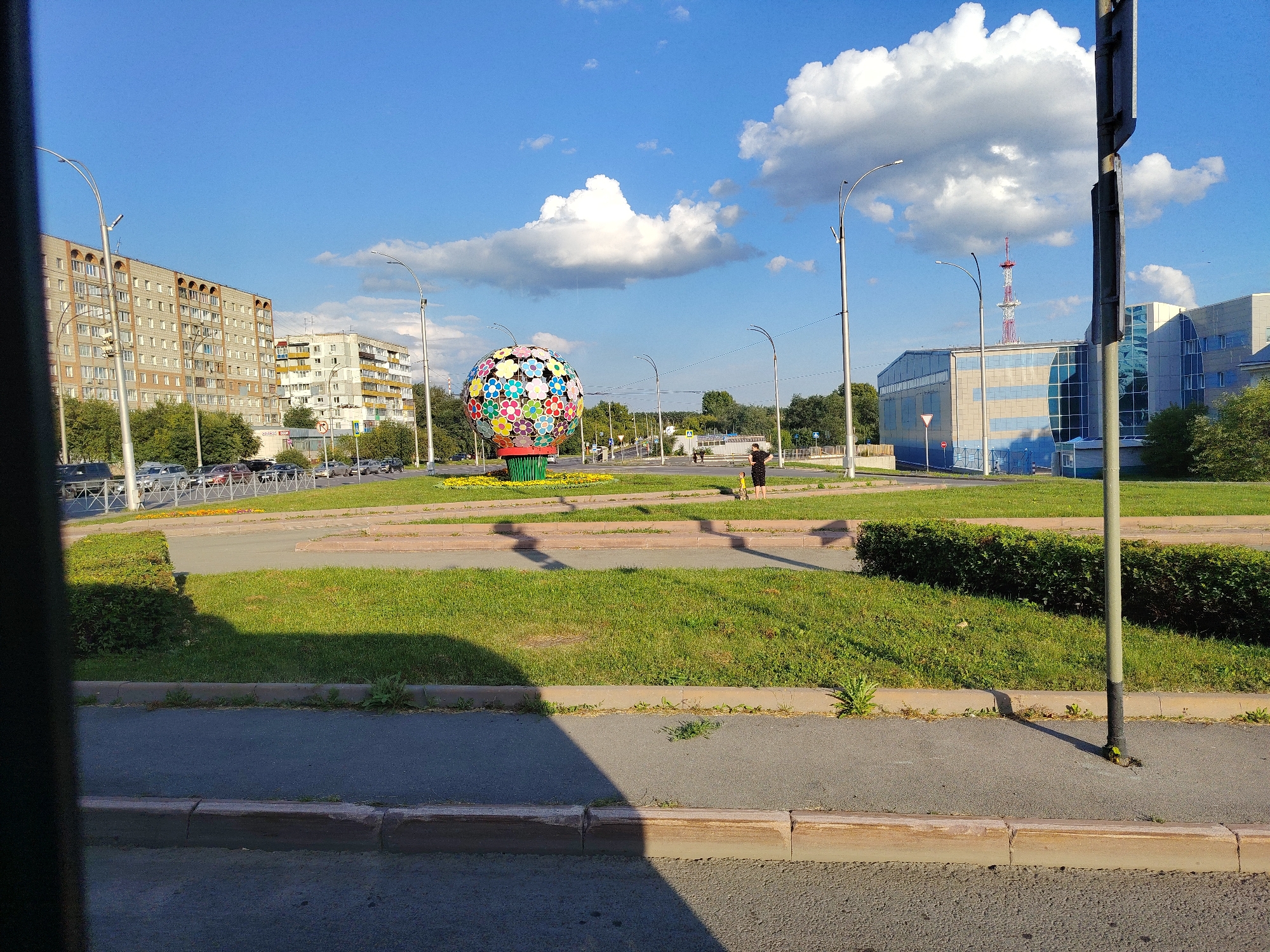 This image depicts a vibrant, spherical sculpture adorned with numerous colorful flower motifs, situated in a public space in Kemerovo, Russia. The sculpture is centrally located on a grassy island, surrounded by a paved pathway and a road with parked cars. In the background, there are multi-story residential buildings and a modern, blue-and-white office building with a radio tower visible behind it. The sky is a clear blue with scattered white clouds, suggesting fair weather. A few people are visible in the distance, including a woman and a child near the sculpture. The scene appears to be a typical urban environment on a sunny day.