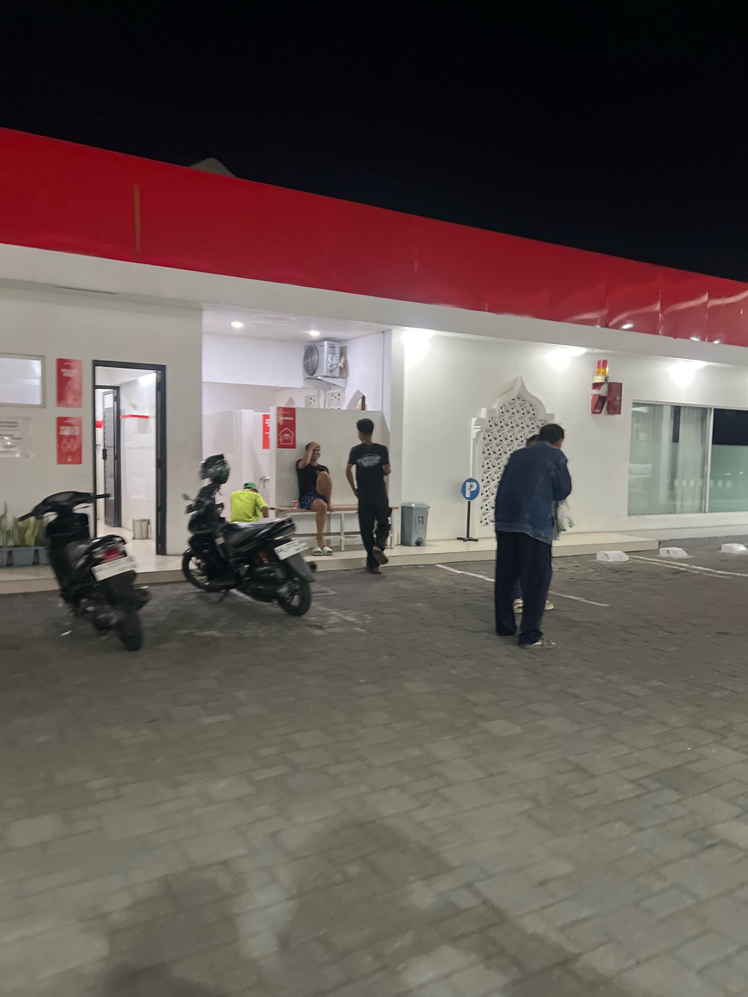 The image captures an outdoor scene at night, likely in Magelang, Indonesia, possibly in front of a commercial establishment with a red and white facade. In the foreground, two motorcycles are parked on a paved surface composed of interlocking bricks. One motorcycle, black, is positioned closer to the left side of the frame, while another, also black, is in the middle ground.
In the middle ground, several individuals are present. Two men stand near a doorway on the left, one of whom appears to be interacting with a woman sitting on a bench inside the building. Further to the right, another man, wearing a dark shirt and pants, walks away from the camera. Next to him, a person in a denim jacket and dark pants is facing away from the camera, holding what appears to be a bag.
The building itself features a white exterior with a prominent red band across the top. The interior visible through the open doorway shows white walls and what might be restroom facilities. Decorative white architectural elements are present on the exterior wall to the right. A blue parking sign with a white 'P' is visible near the person in the denim jacket. The lighting suggests artificial illumination from the building and its surroundings. The background is dark, indicating nighttime.
