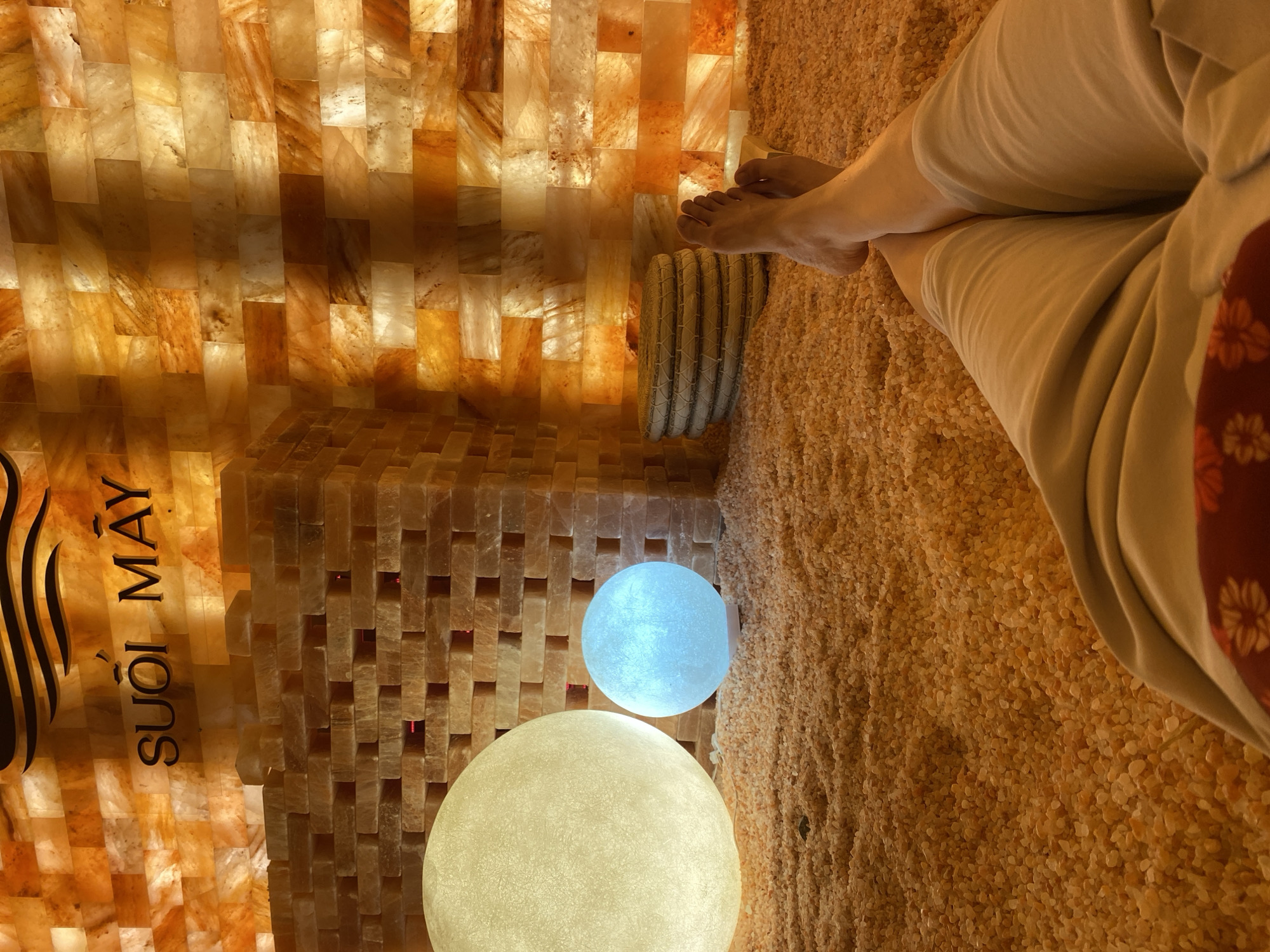 The image captures an indoor salt room, likely a spa or wellness facility, at Suối Mây in Da Lat, Vietnam. The walls are constructed from illuminated salt blocks in various shades of orange, yellow, and white, creating a warm and ambient glow. In the foreground, a person's bare legs and feet are visible, relaxed and positioned on a bed of small, light-orange pebbles that cover the floor. Two large, spherical lamps are present in the midground: one emits a soft yellow light, while the other glows with a blue hue. A stack of circular, woven mats rests beside the blue lamp. The overall impression is one of tranquility and relaxation within a uniquely designed space.