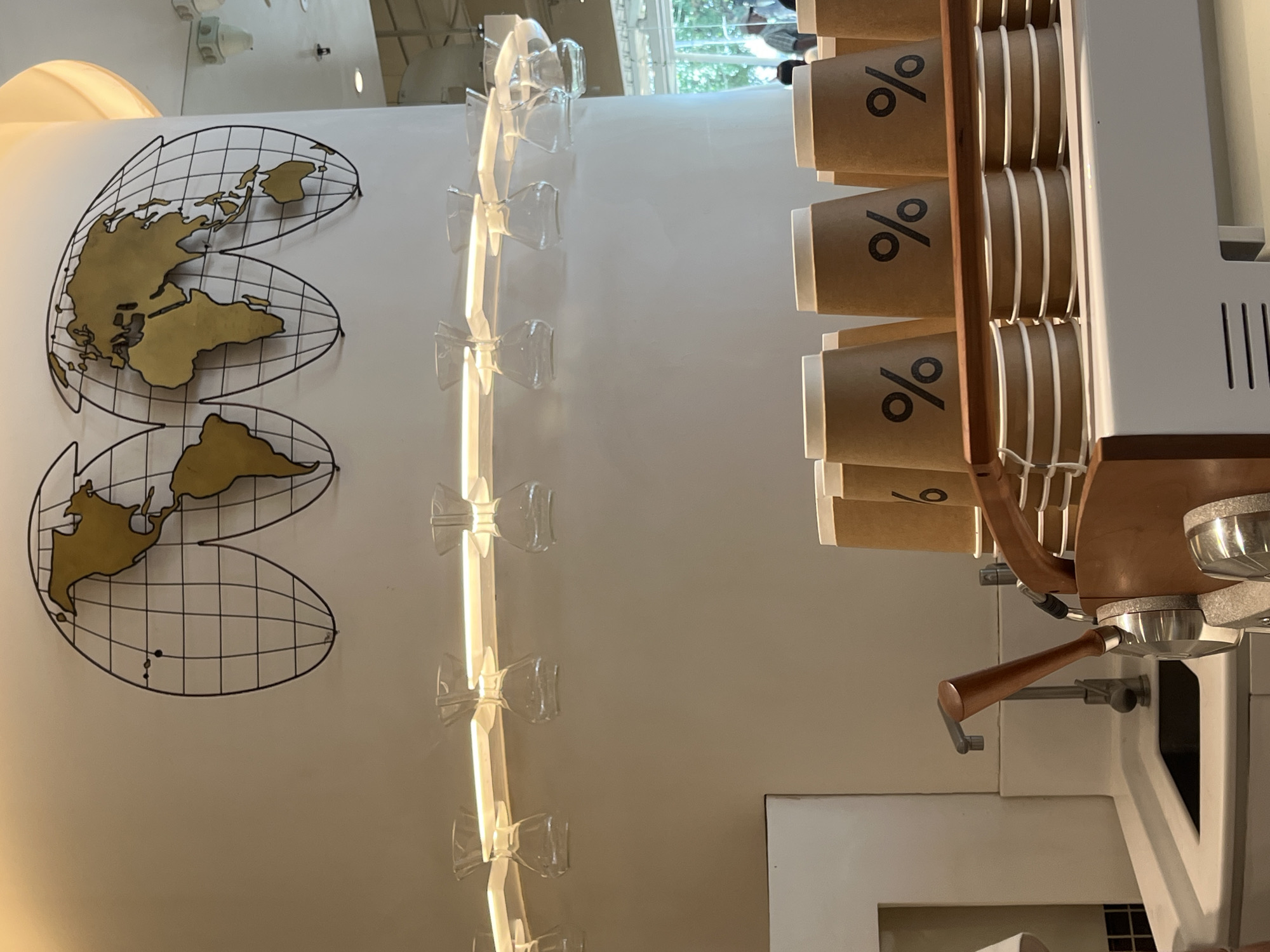 This image depicts the interior of a coffee shop, likely a high-end establishment, with a focus on coffee preparation and presentation. The background features a stylized world map as wall art, suggesting an international theme or brand. Below the map, a row of small glass pour-over coffee drippers is suspended from a light fixture, indicating a focus on manual brewing methods. In the foreground, a stack of brown paper coffee cups with the percentage symbol logo is neatly arranged, ready for use. The scene includes a stainless steel sink and faucet, a coffee machine handle, and a wooden rack holding the cups. The overall aesthetic is clean, modern, and minimalist, with warm lighting creating an inviting atmosphere. The presence of outdoor greenery visible through a window in the background suggests the coffee shop is located in an urban environment. While the specific location within Kuta, Indonesia, is not explicitly identifiable from the image alone, the style and branding are consistent with modern coffee shops commonly found in tourist-friendly areas like Kuta. No people are clearly visible in the immediate foreground, though figures can be faintly discerned in the background through the window.