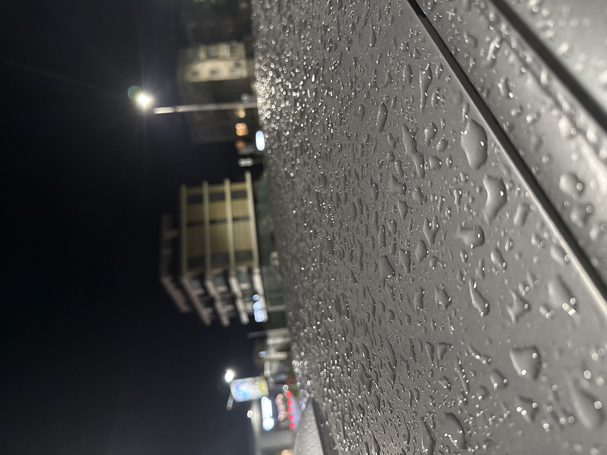 The image is a close-up view of a wet surface, likely metal, covered in numerous water droplets. The droplets vary in size, reflecting light and creating a glistening effect. The angle is slightly skewed.
In the background, out of focus, are buildings that appear to be located in a city. There are building with multiple floors, street lights, and signs with illuminated lettering. The blurry background suggests the image was taken at night. The location is possibly Plovdiv, Bulgaria, given the prompt. However, without specific architectural or signage details, a precise identification of the exact location is impossible.