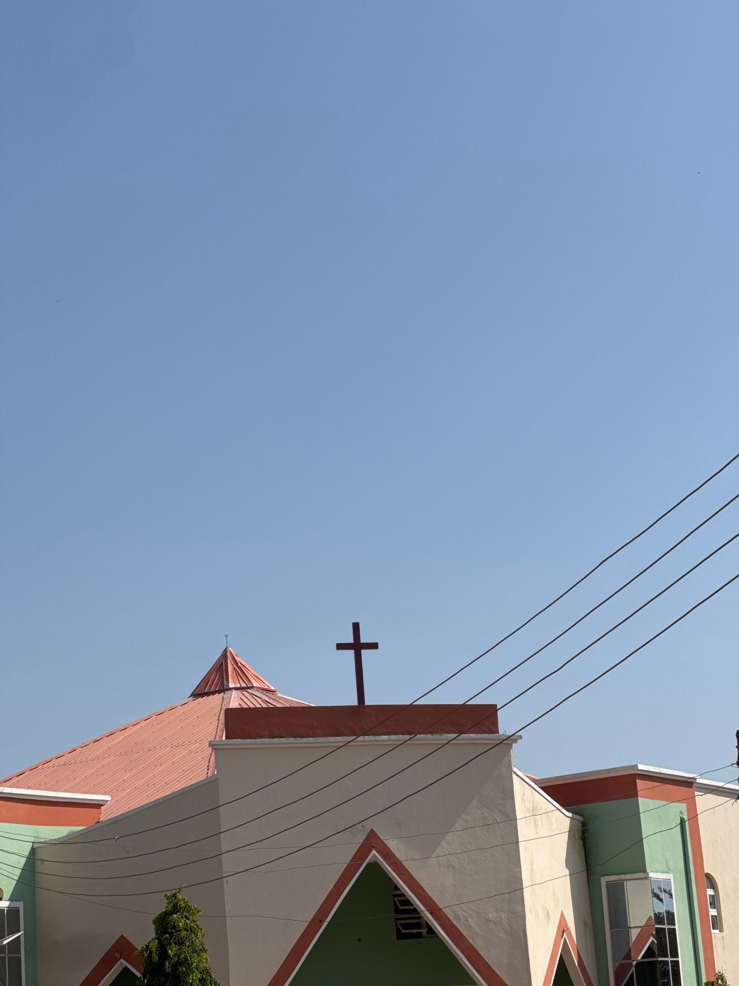The image shows the upper portion of a church building against a clear blue sky. The church has a red, conical roof with a cross on top. The building itself is painted in cream, green, and red. Several power lines are visible in the foreground, crossing the sky and the building. A small tree is visible in the lower left of the frame. There are no people visible in the image.