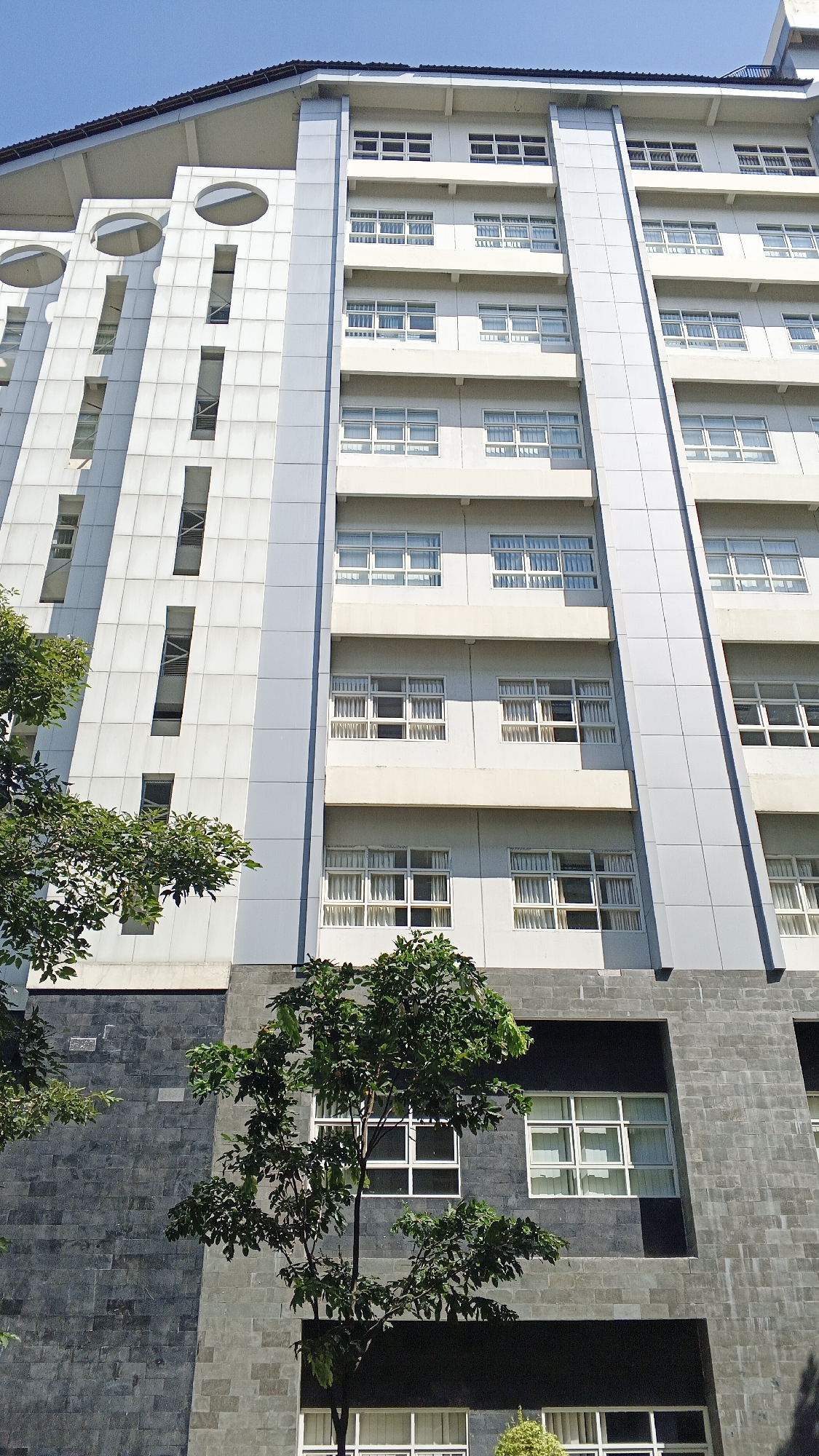 This is a medium-angle, exterior, full-shot photograph of a multi-story building with a light gray facade and a darker gray, stone-like base. The building is tall and appears to be an institutional or commercial structure. The sky is a clear, bright blue.
The building's facade is divided into horizontal sections, with alternating lighter and darker gray bands. The upper floors feature numerous windows, arranged in rows and columns. Many of these windows are large and have white frames, with sheer white curtains visible inside. Some windows are divided into multiple panes. On the left side of the building, there are several vertical, rectangular openings with recessed windows or darker interiors, and some larger circular indentations.
In the foreground, on the left and bottom of the image, are several leafy green trees. The tree on the left is closer to the building and partially obscures some of the lower windows. The tree in the bottom center is smaller and has denser foliage. The base of the building is clad in dark gray, rectangular stone tiles, which have a textured appearance.
The scene is well-lit, suggesting it was taken during daylight hours, likely in the morning or afternoon due to the clear sky and shadows. No people are visible in the image. The overall impression is one of a modern, functional building set against a natural backdrop of trees. This building is located in the city of Banguntapan, Indonesia.