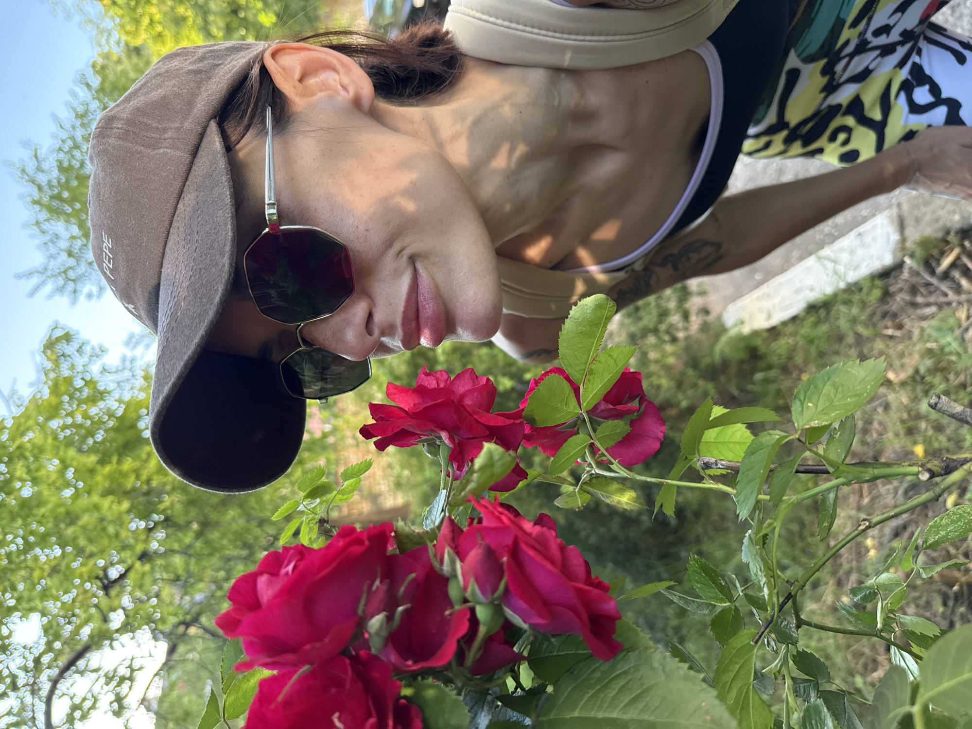 A woman in a brown cap and sunglasses is looking at a bouquet of red roses. The photograph appears to have been taken from a low angle, with the roses prominently in the foreground and the woman leaning over them. The background shows trees and sky, indicating an outdoor setting. There is no clear information in the image to confirm that the location is in the city of Pleven, Bulgaria.