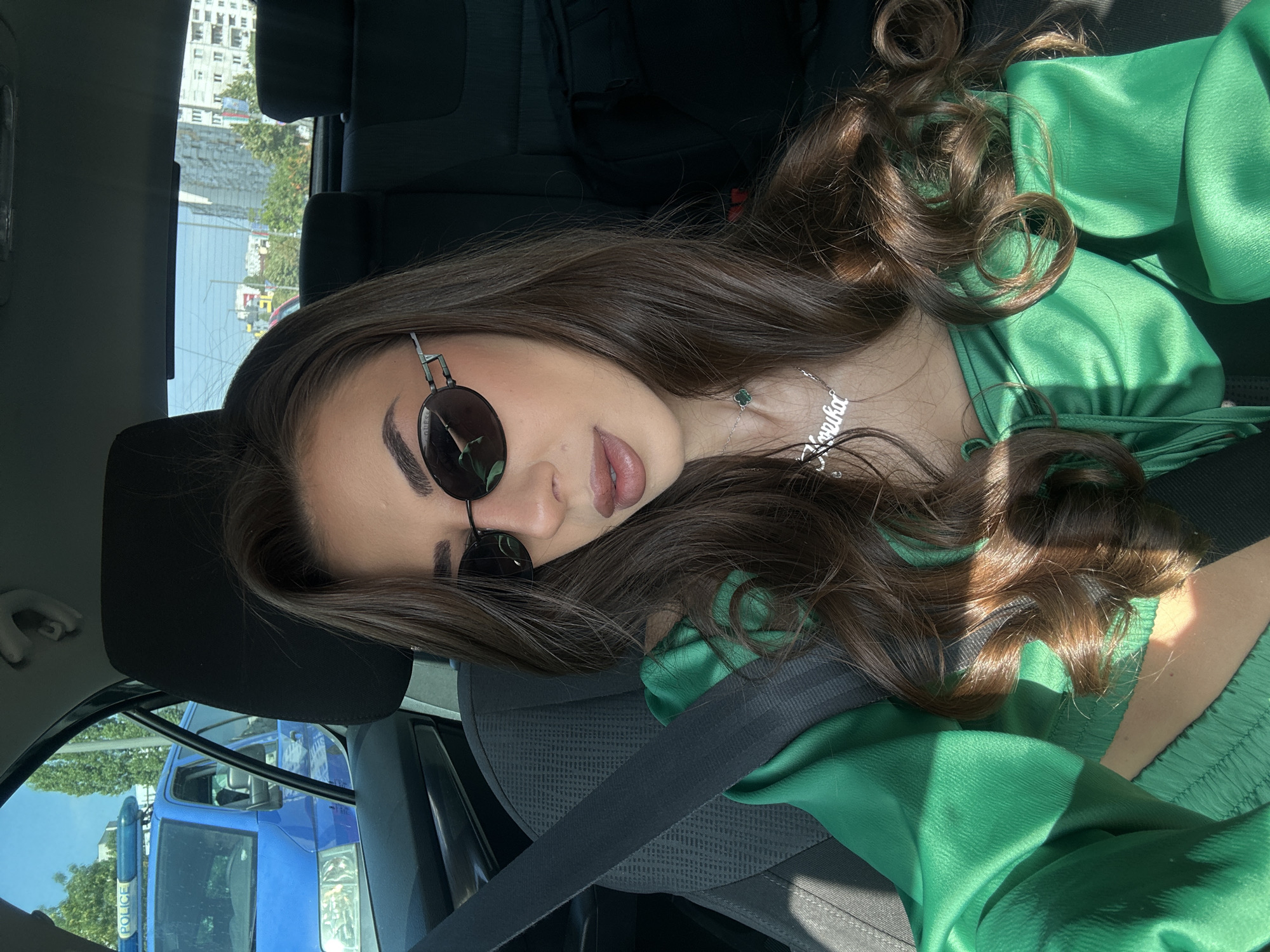 In Sofia, Bulgaria, a woman is seated in the driver's seat of a car. She is wearing a green top, sunglasses, and a necklace. Through the car window, a blue police van and other vehicles are visible, along with buildings and trees, suggesting an urban setting. The woman's body is turned slightly towards the camera, and her hair is down. The car's interior, including a seatbelt, is also in view.