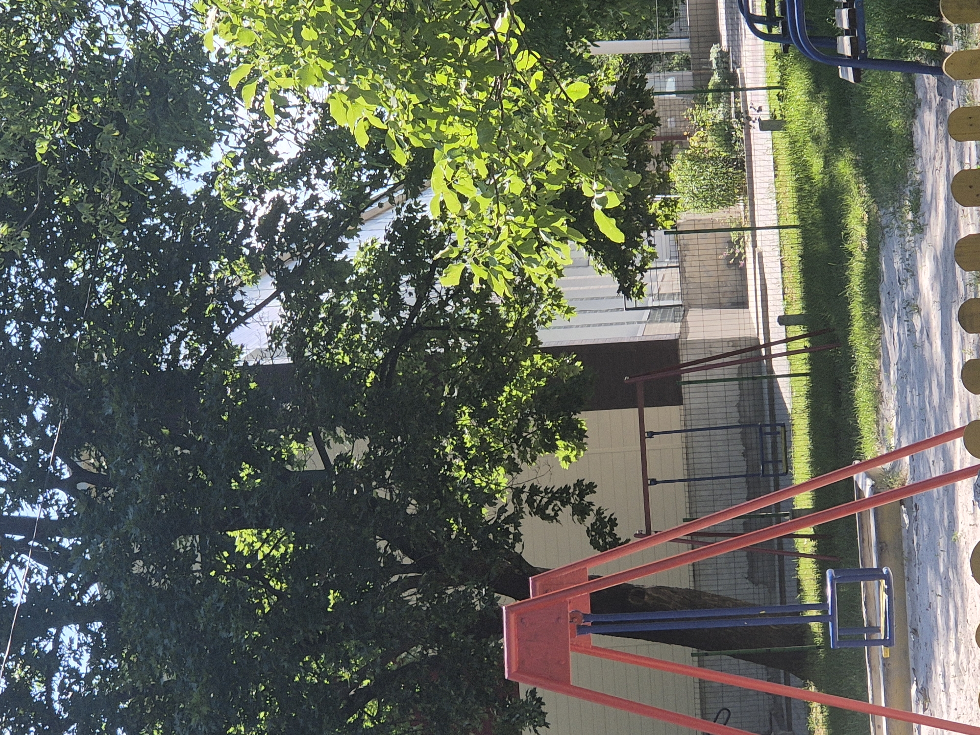 The image shows a playground area with a red A-frame swing set in the foreground. A blue swing seat hangs from the swing set. Behind the swing set, a chain-link fence encloses a grassy area with more playground equipment, including another swing set with blue seats. Trees with dense green foliage are visible in the background, and a building with light-colored siding is partially obscured by the trees. The ground in the foreground is covered with sand and small yellow decorative elements. The scene appears to be in an outdoor public space on a sunny day.