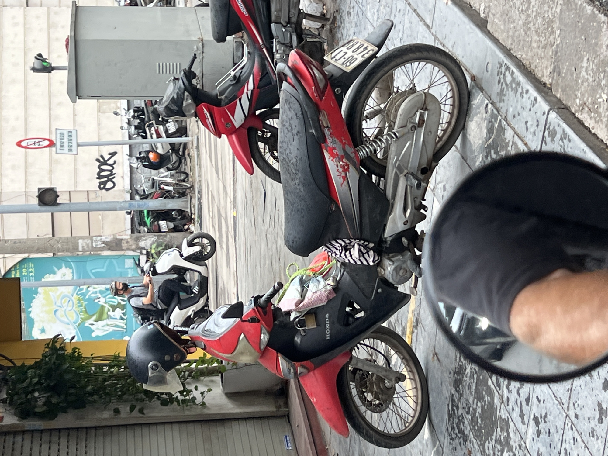 The image captures a street scene in Hanoi, Vietnam, focusing on a row of parked motorcycles. In the foreground, a red and black motorcycle is prominently displayed, with its front wheel, handlebars, and part of the engine visible. A black helmet rests on the handlebars. To the right, the rear section of another red motorcycle is seen. In the background, a person wearing a helmet and a face mask is riding a white motorcycle. The street is paved with concrete, and there are various signs and a traffic light visible. Buildings line the street, and some greenery is present on the left side of the image. The overall impression is one of a typical urban environment in Hanoi, characterized by its busy street life and prevalence of motorcycles.