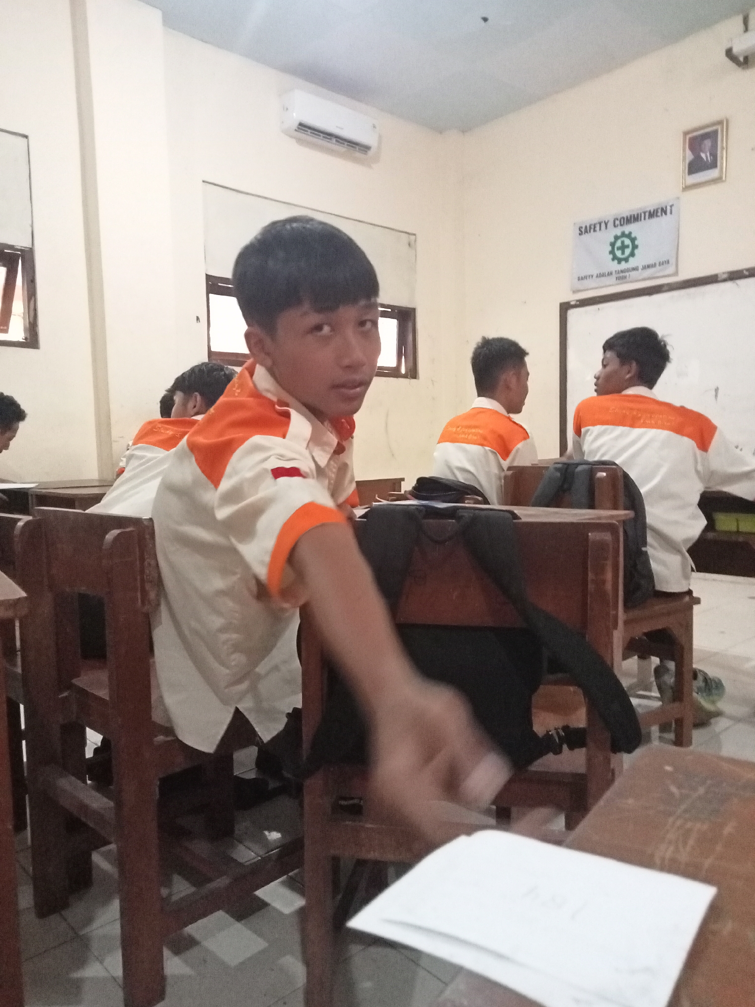 In a classroom in Secang, Indonesia, a group of male students are seated at wooden desks. The student in the foreground, facing the camera, wears a white shirt with orange accents on the collar and sleeves. His hand is extended towards the camera, and he is looking directly at it. Behind him, other students are also visible, some wearing similar uniforms. In the background, a whiteboard displays a "Safety Commitment" sign with a green safety symbol. An air conditioning unit is mounted on the wall. The room appears to be illuminated by overhead lighting and natural light filtering through windows.