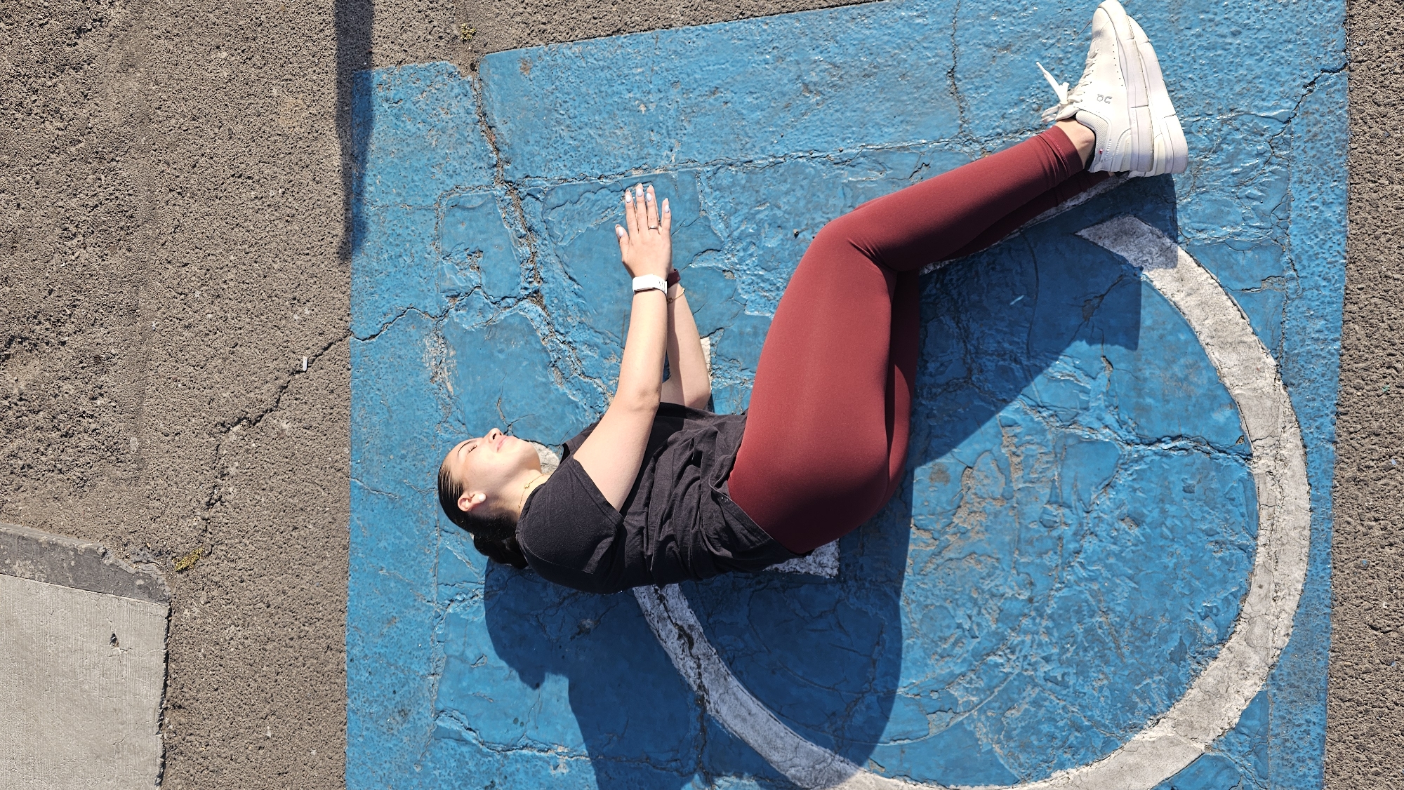 A woman lies on her back on a blue painted parking space. The parking space has a white painted wheel chair symbol, which is cracked. The woman has a black T shirt and dark red leggings, and white athletic shoes. She is wearing a white wrist watch on her left wrist and two rings on her left hand. Her arms are lifted upward, and her palms are facing each other. The painted parking space is bordered by asphalt concrete. The scene is illuminated by bright sunlight. The scene is in Puebla, Mexico.