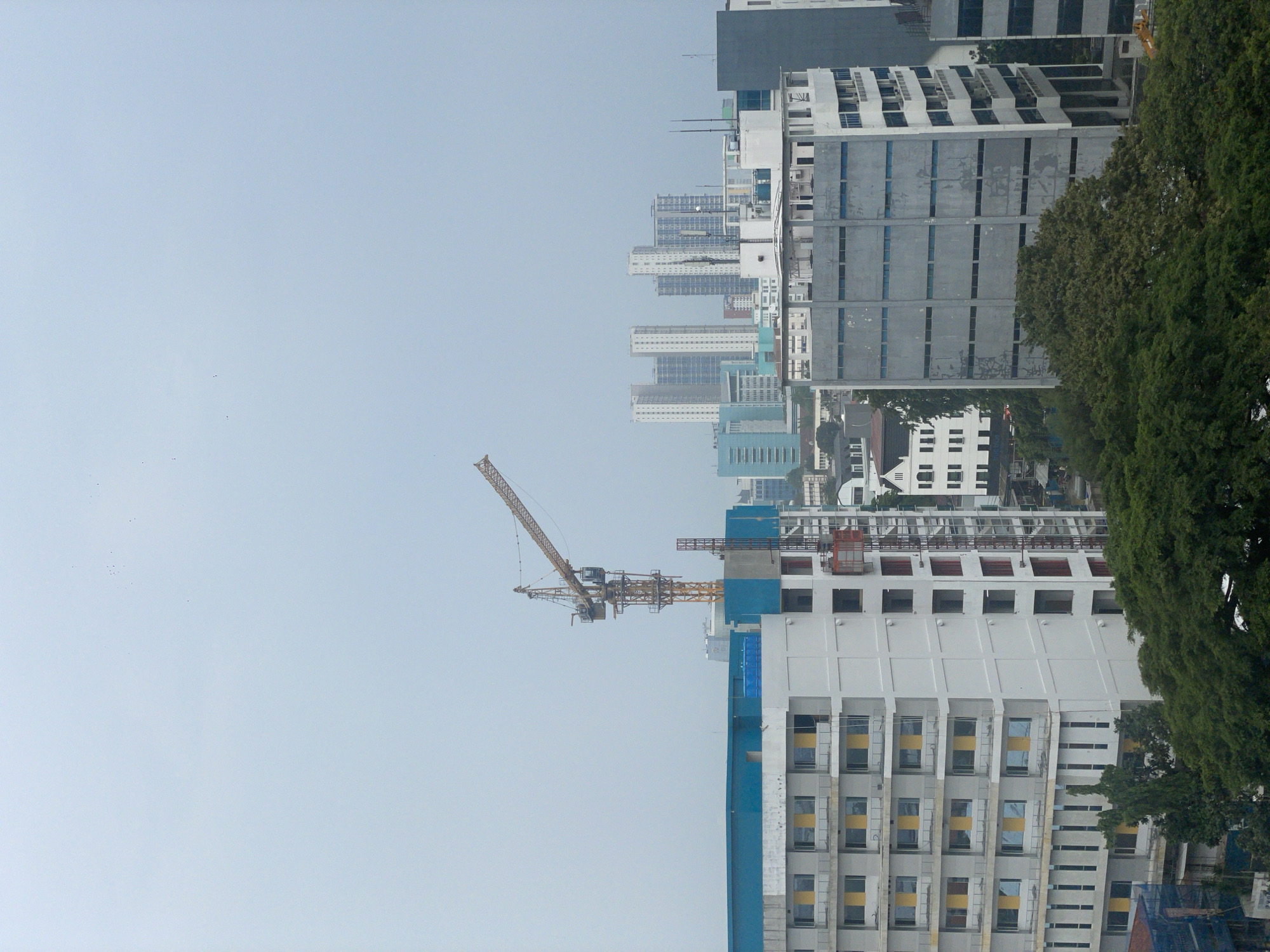 This image shows a cityscape, likely in Central Jakarta, Indonesia, dominated by a construction site in the foreground. A tall building under construction is partially visible, with a yellow crane extending into the sky. The building has a white and blue facade with rows of windows.
Behind the construction site, the city skyline includes various mid-rise and high-rise buildings, some covered in gray siding. Trees frame the edges of the image on the right. The sky is overcast, creating a muted color palette throughout the scene. No people are visible.