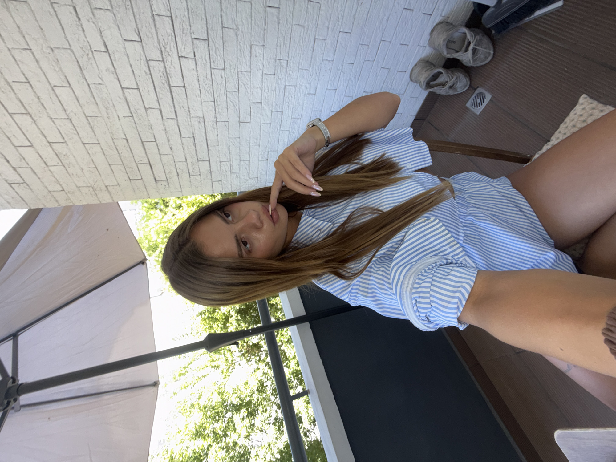 An adult female with long, brown hair is sitting on a chair on a balcony. She is wearing a blue and white striped shirt. Her left hand is touching her chin. In the background, there is a white brick wall, green trees, and an umbrella. Two pairs of grey shoes are visible on the floor. The location is identified as Plovdiv, Bulgaria.