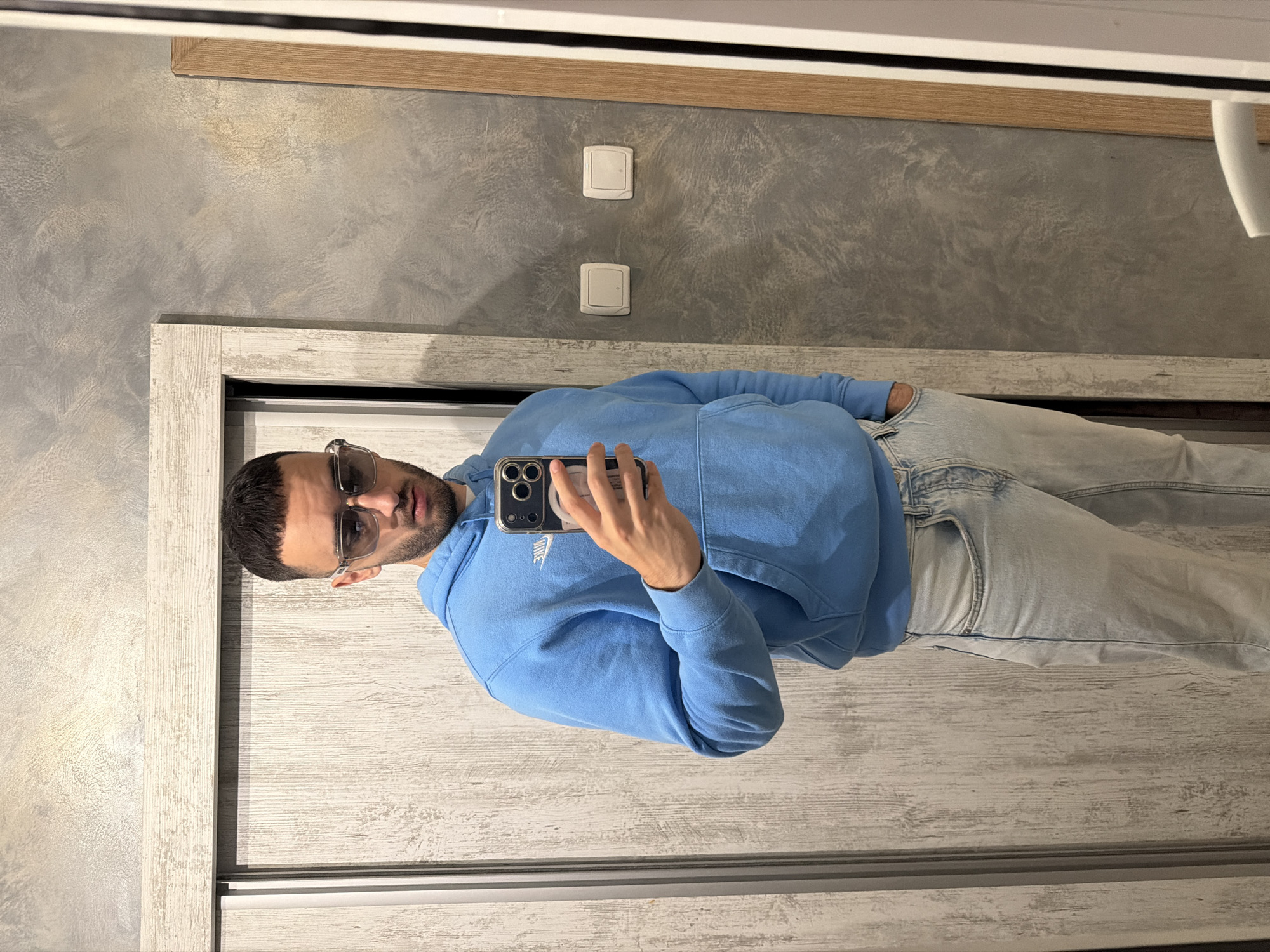 A man with short dark hair and a beard stands in front of a mirror, holding his phone to take a selfie. He wears sunglasses, a light blue Nike hoodie, and light-wash jeans. The background includes a textured gray wall with two light switches and a wooden frame. The man appears to be indoors, possibly in a closet or wardrobe area. It is not possible to determine the location in the city of Plovdiv, Bulgaria, from the image.