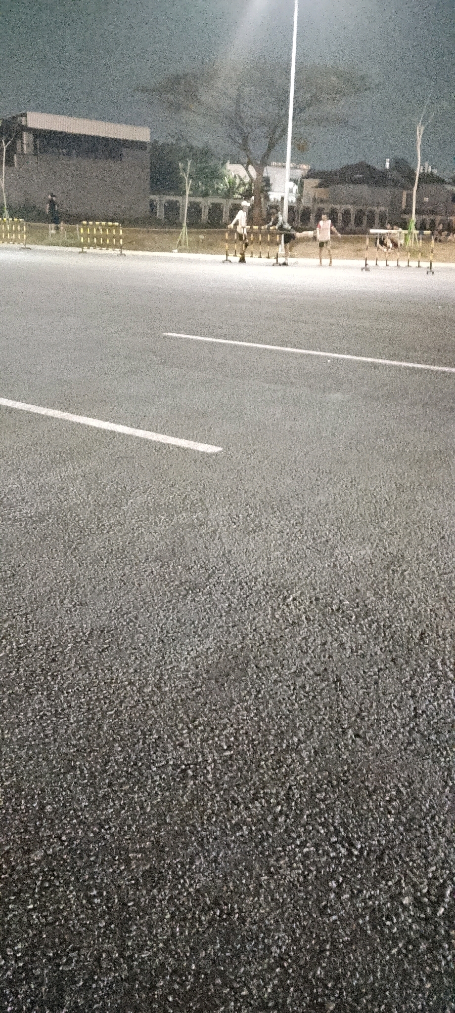 A low-angle, grainy, nighttime photograph captures an outdoor scene in Gunung Putri, Indonesia, likely a sports or training facility. The foreground is dominated by the textured asphalt of a road or track, marked with two parallel white lines. In the mid-ground, a group of four individuals, appearing to be young males, are engaged in some form of physical activity, possibly an obstacle course, as they are positioned around a series of low hurdles. They are all dressed in athletic attire. Behind them, a long, low building with a light-colored facade is visible, suggesting it might be a barracks or administrative building. Yellow and black striped barricades line the edge of the paved area in front of the building. The background features trees and residential-style buildings under a dark, overcast sky, illuminated by a bright overhead light, likely a streetlamp. The overall impression is one of a dimly lit, functional outdoor space.