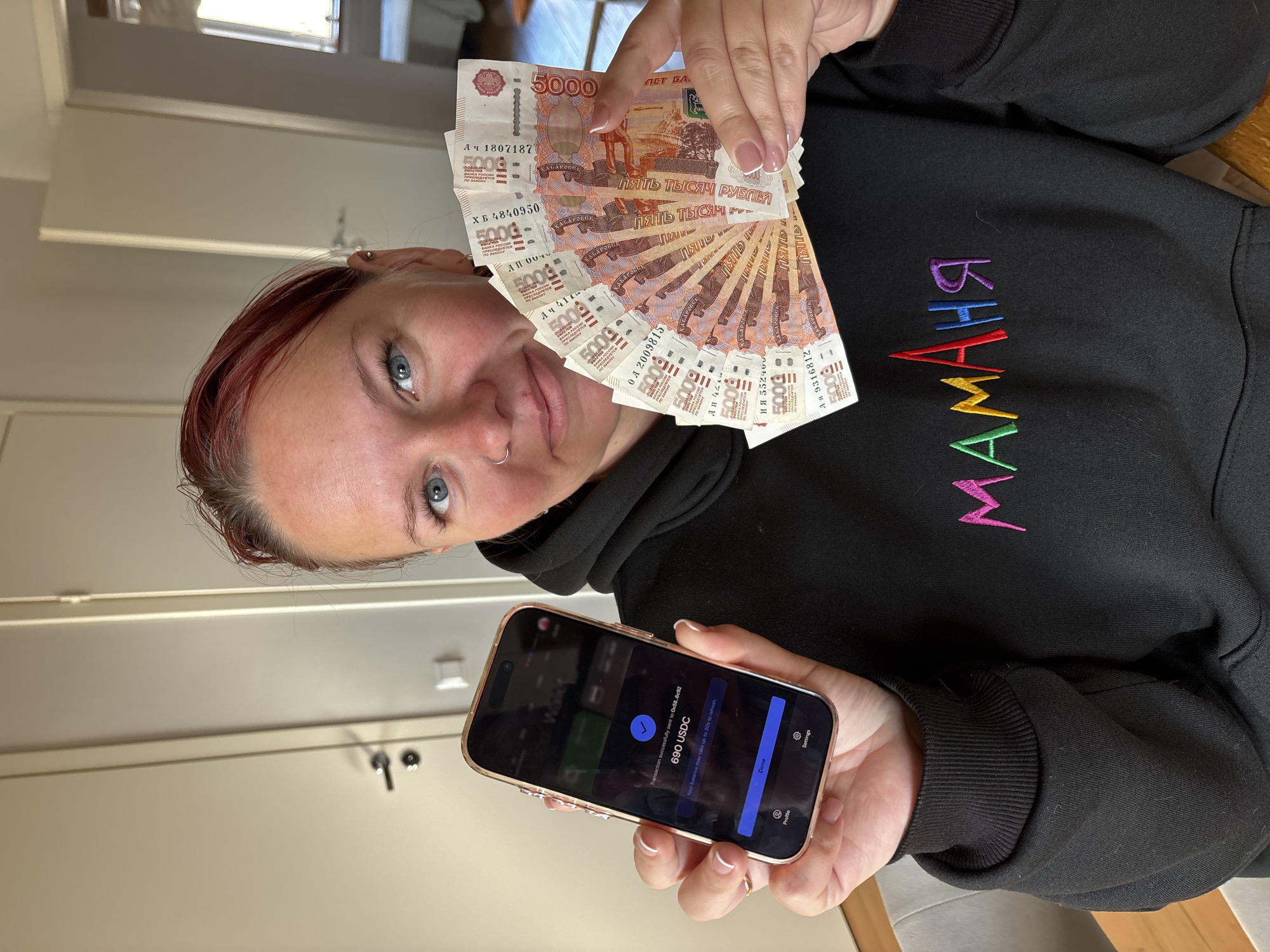 An indoor, portrait-style photograph, taken in Chaikovsky, Russia, features a woman looking directly at the camera. She has reddish-brown hair, blue eyes, and a nose ring, with a neutral to slightly positive expression. Her nails are styled with a French manicure.
In her right hand, she holds a fan of multiple 5000 Russian ruble banknotes, clearly displaying the "5000", "ПЯТЬ ТЫСЯЧ РУБЛЕЙ" (Five Thousand Rubles), and "ХАБАРОВСК" (Khabarovsk) text, along with various serial numbers such as "АЧ 1807187", "ХБ 4840950", and "АЯ 9316812".
In her left hand, she holds a smartphone, likely an iPhone in a rose gold or gold case, displaying a cryptocurrency transaction. The phone screen shows a "Transaction successfully sent to 0x58...6c92" message and "690 USDC", with "Done", "Settings", and "Profile" options visible.
The woman is wearing a black hooded sweatshirt with colorful embroidered Russian text "МАМАНЯ" (MAMANYA), meaning "Mommy" or "Mama," in purple, blue, green, yellow, and red threads. The background reveals a simple, light-colored room interior, suggesting a residential setting during daylight hours. The image juxtaposes physical cash with a digital currency transaction.
