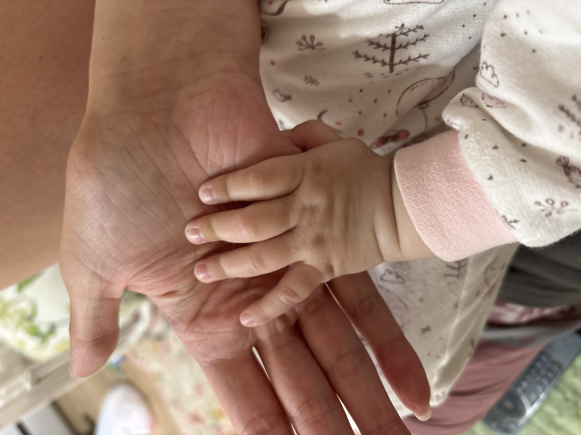 This close-up image captures a poignant moment of connection between an adult and a baby. The central focus is on a large, open adult hand, positioned on the left side of the frame with its palm facing upwards. A small baby's hand rests gently upon the adult's palm, with its tiny fingers curled, emphasizing the size contrast between the two. Both hands appear light-skinned.
The baby is dressed in a light-colored, long-sleeved garment, possibly a bodysuit or sleepsuit, adorned with a whimsical pattern of small brown-toned trees, stars, and other abstract shapes like clouds or animals. The cuff of the baby's sleeve is a soft pink.
The background elements are largely out of focus, with a shallow depth of field drawing attention to the hands. On the far left, a blurred portion of an adult's skin is visible. The lower left shows a soft, out-of-focus patterned fabric and a white shoe or slipper. To the right of the baby's arm, dark fabric and a faint outline of what appears to be a remote control or similar electronic device can be seen, indicating an indoor setting.
The image conveys a strong sense of tenderness, warmth, and a loving bond between the two individuals. There is no visible text in the image. The scene is set indoors in Sochi, Russia.