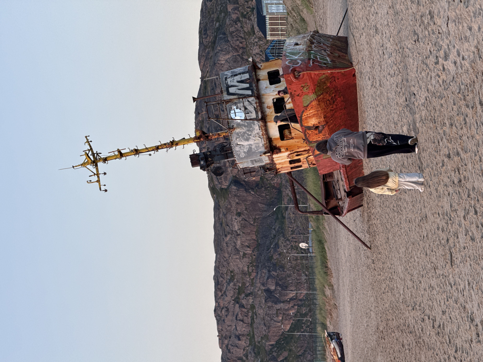 A rusted shipwreck lies beached on a sandy shore in Teriberka, Russia, under a clear sky. The ship, tilted on its side, is adorned with graffiti. Two people, a woman in a grey hoodie and a young girl in a yellow jacket, walk away from the shipwreck across the sand. In the background, rocky hills rise behind a grassy area with streetlights and a fence. Further in the distance, modern buildings with glass facades are visible. Another smaller, partially visible shipwreck lies to the left of the main subject.