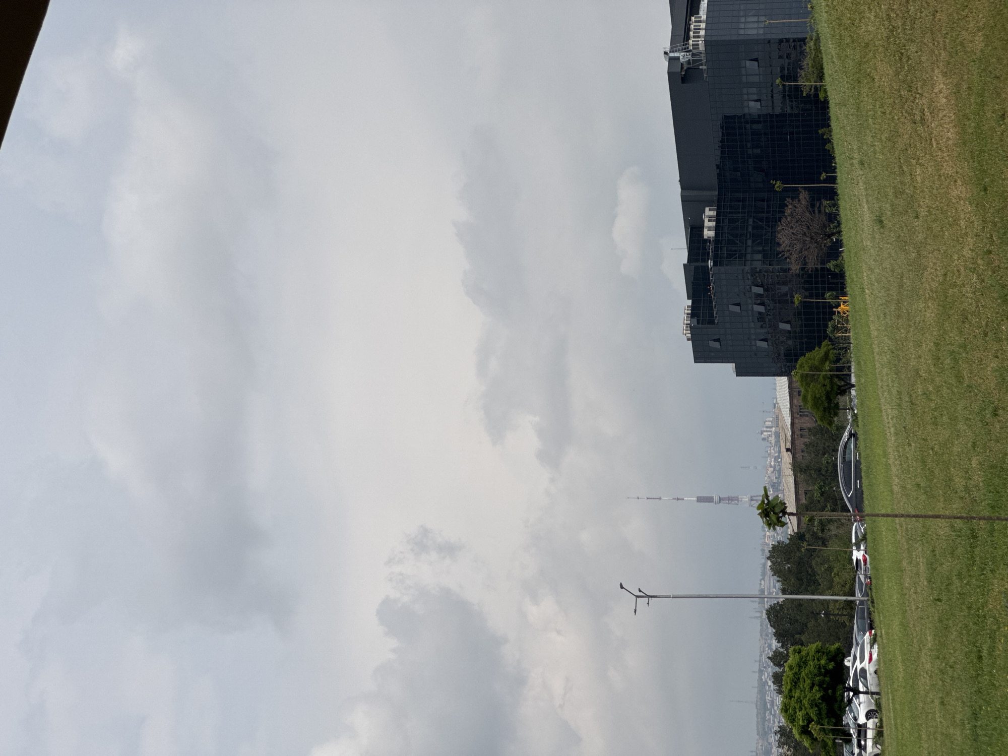 The image shows a partial view of Yerevan, Armenia, under an overcast sky. A modern, dark-glass building stands adjacent to a green lawn. Further in the distance, the Yerevan TV Tower is visible, rising above the cityscape. A row of parked cars lines the edge of the lawn. Trees and other vegetation are interspersed among the buildings. A streetlight pole is present in the mid-ground.