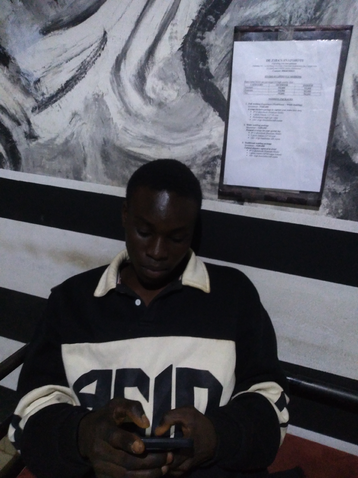 A young man is seated in a dimly lit indoor space in the city of Unknown, Unknown. He has dark skin and short, dark hair, and is looking down intently at a dark-colored smartphone held horizontally in both hands. He is wearing a long-sleeved polo shirt featuring a black upper section, a white middle section with dark, partially legible lettering (possibly "SNAPS"), and black sleeves with white-striped cuffs. His collar is off-white.
Behind him, a wall is adorned with abstract black and white/gray brushstroke art depicting swirling and wavy patterns. To the right, a rectangular framed document is mounted on the wall. The white document, partially legible, is titled "DR ZIHAN SNAPSOTS" and lists details for "FAMILY & LIFESTYLE SESSIONS" and "WEDDING PACKAGES" with associated prices, indicating the location is likely a photography studio or related commercial establishment. Below the wall art, a black structure with two horizontal white stripes is visible, possibly a part of a bench, a railing, or a decorative element. A reddish-brown floor or surface is partially visible in the lower right corner.