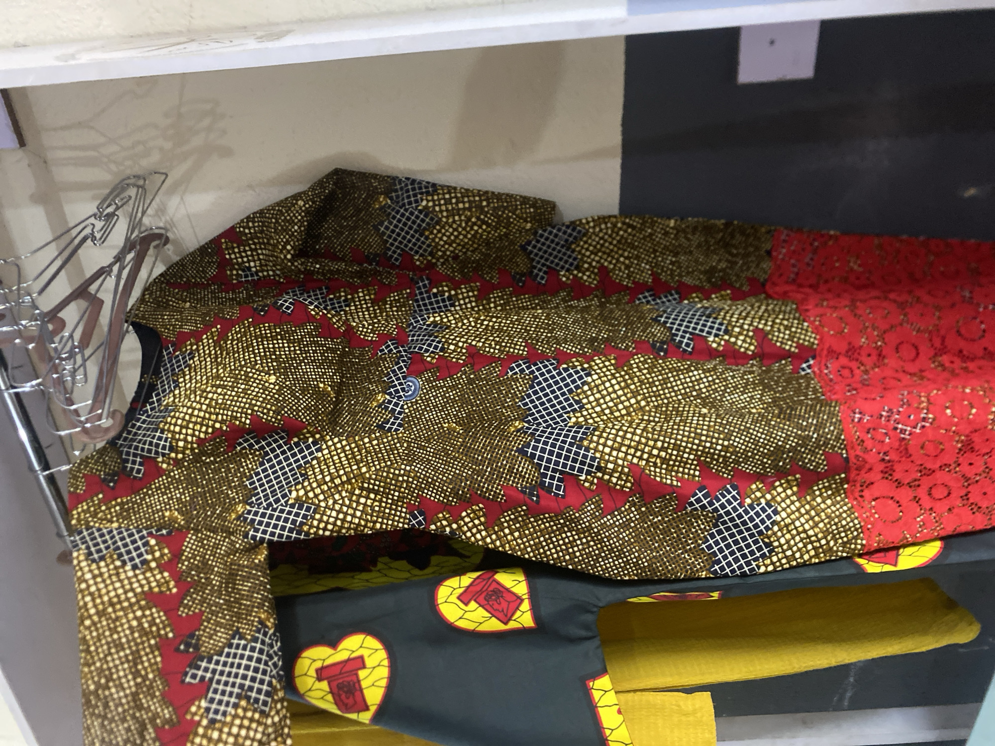 A colorful, traditional African dress hangs on a clothes hanger inside a closet. The dress is made of a vibrant ankara print fabric with a pattern of gold, red, and black. It has a lace trim at the hem. To the left of the dress, another garment is partially visible, appearing to be yellow with a dark gray fabric overlay featuring yellow and red heart-shaped motifs. The closet interior features a white wall, a dark gray lower wall, and a clothes rail. The image was taken in Bukuru, Nigeria.