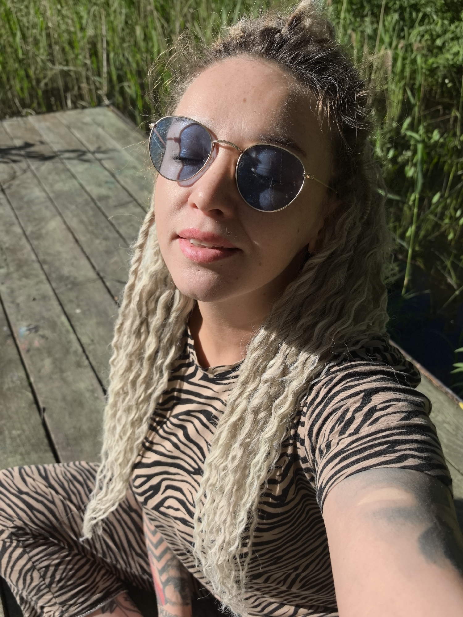 A woman with fair skin, blonde dreadlocks pulled into a bun, and wearing round sunglasses is seated on a wooden dock. She is wearing a brown and black zebra print top and pants. Her eyes are closed and she has pink lipstick on. Tattoos are visible on her left arm and leg. The background shows green reeds and a body of water. The location is Noviy, Russia.