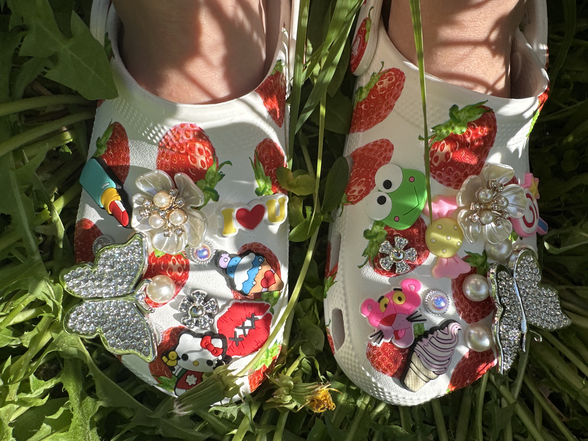 Two feet wearing white Crocs with strawberry patterns and various decorative charms stand in green grass. The Crocs are adorned with charms including butterflies, flowers, lipstick, an "I <3 U" symbol, cartoon characters (Hello Kitty, Pink Panther, a green frog), sweets (ice cream), and pearls. The grass is green and dense, with some dandelions. The photo is taken outdoors in natural sunlight. There is no indication that this image was taken in Nizhny Novgorod, Russia.