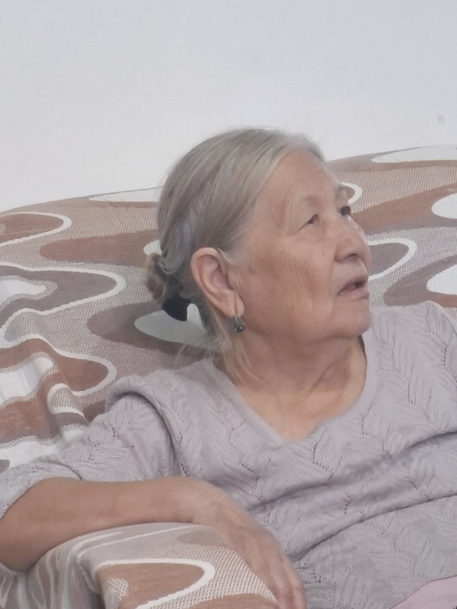 A medium shot, eye-level, indoor portrait features an elderly East Asian woman with grey hair pulled into a bun and a single dark earring in her left ear. She is seated on a couch with a patterned throw, wearing a light gray knitted sweater. Her head is turned to her right, and her gaze is directed upwards and to her right, with her mouth slightly open. The background is a plain white wall. The scene is devoid of any identifying landmarks or features that would indicate its location within Almaty, Kazakhstan. The focus is entirely on the woman's face and upper body.