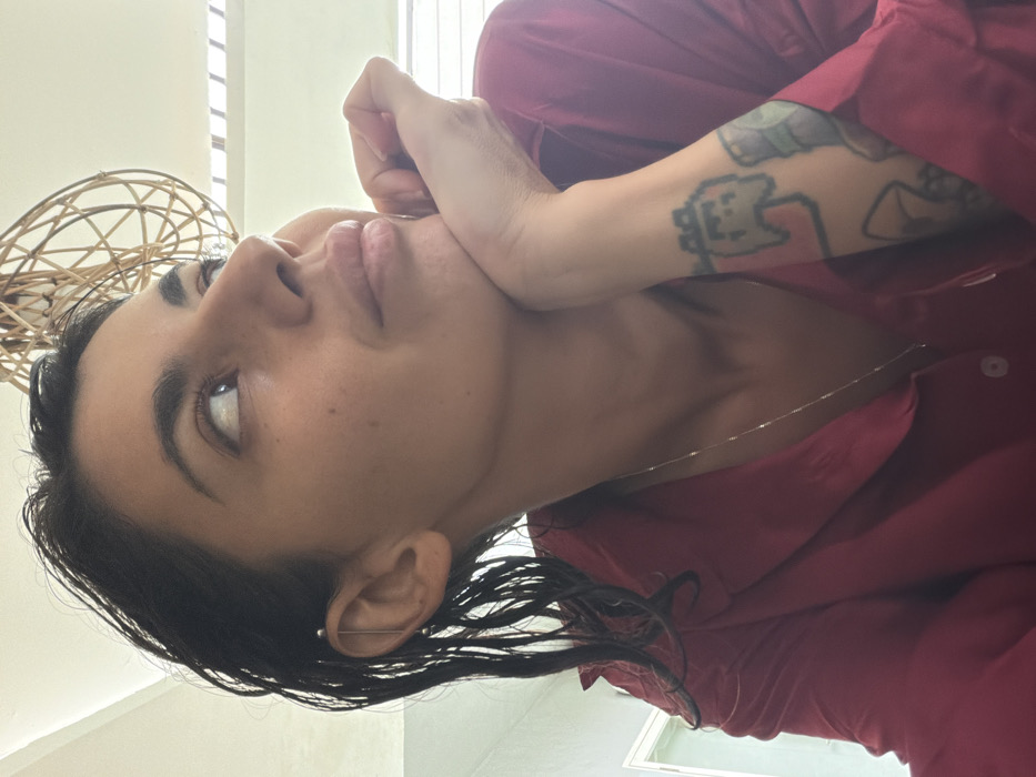 The image displays an adult female indoors, located in Nusa Dua, Indonesia. She is seen from the chest up, with her left hand positioned under her chin, supporting her head. Her dark hair appears wet and is styled away from her face. A silver-toned industrial piercing is visible in her left ear. She wears a red collared shirt and a thin silver-colored chain necklace. Her left forearm features multiple tattoos, including a prominent light-green pixelated ghost-like figure. The subject's gaze is directed upwards and to the right, beyond the frame. The background consists of a light-colored ceiling from which a woven, spherical decorative object or light fixture is partially suspended. A portion of a wall or doorframe is visible on the far left.
