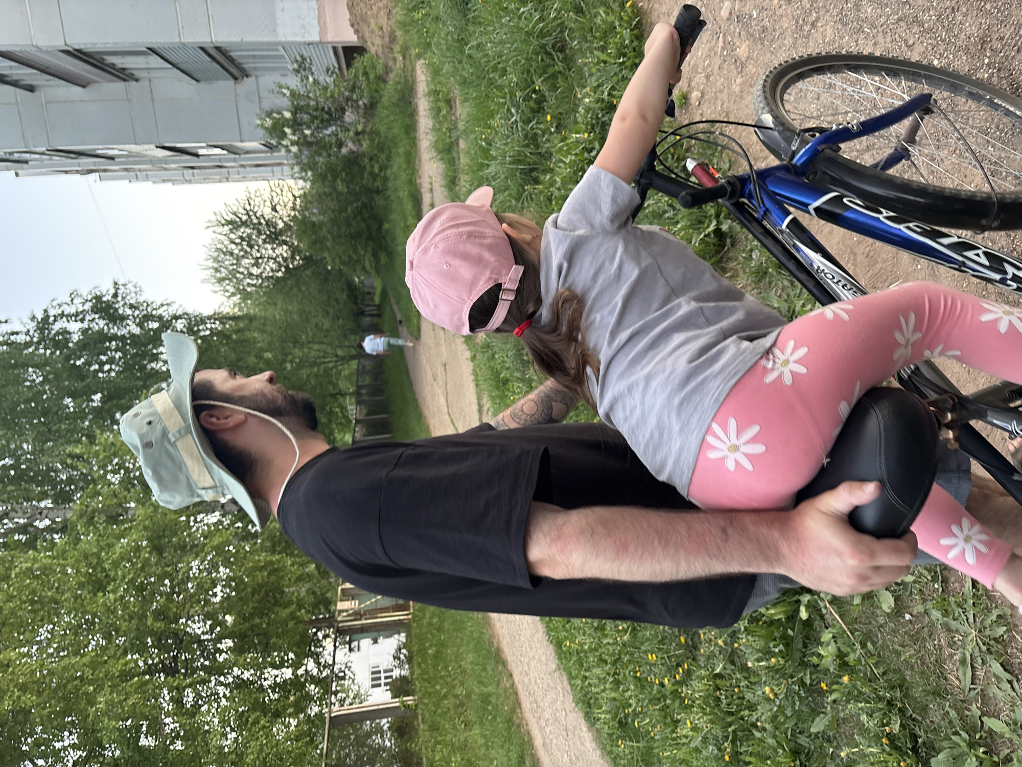 A father, wearing a light blue bucket hat and black t-shirt, supports his young daughter, who is wearing a pink baseball cap, gray t-shirt, and pink leggings with white flower patterns, as she sits on a blue and black bicycle. The scene appears to be in a grassy area in Chaikovsky, Russia, with buildings and trees visible in the background. The father has a beard and is looking at his daughter. The daughter is holding the handlebars of the bicycle. There are yellow flowers dotting the grass.