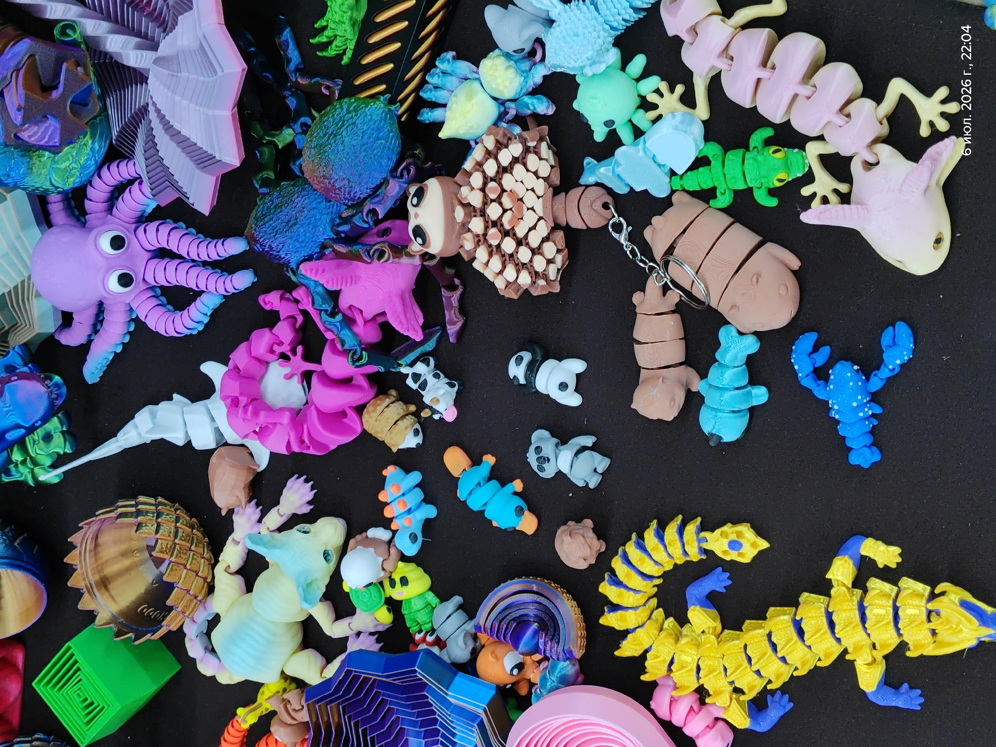 The image shows a flat lay of various 3D printed toys on a dark surface. The toys include an octopus, a dragon-like creature, a segmented alligator, a mushroom-shaped figure, a lobster, a cat, a koala, pandas, and other miscellaneous creatures. The items are arranged in a dense, somewhat chaotic manner, with some overlapping. The lighting suggests an indoor environment. There is no discernible indication of the city of Astana, Kazakhstan, or any urban elements.
