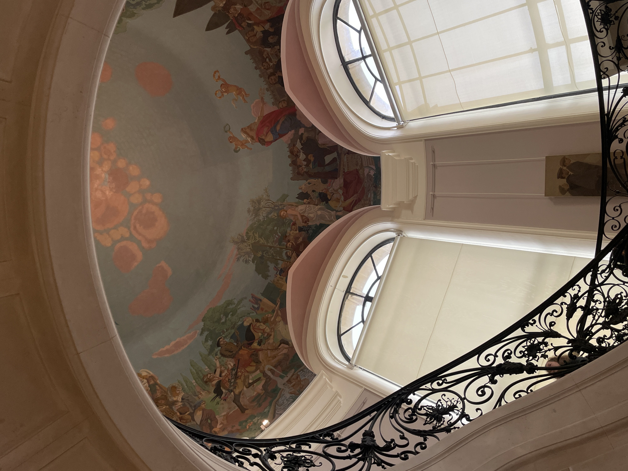 The image provides an upward interior view of the main staircase and domed ceiling within the Petit Palais, located in Paris, France. A grand, curving staircase, defined by its ornate dark wrought-iron railing featuring intricate scrollwork and floral designs, ascends along a light-colored architectural structure.
Above, a large domed ceiling is covered by a detailed fresco painting, consistent with "The Triumph of Paris" by Fernand Cormon. The fresco depicts multiple narrative scenes: one section shows figures on horseback, suggesting a battle or procession, set against a green landscape; another portrays groups of individuals in classical attire, both standing and seated, indicating a gathering or intellectual assembly. Flying cherubs holding wreaths and a central figure draped in red and blue garments are also visible. The fresco's background sky is light blue with diffuse orange and pink cloud-like formations.
Below the frescoed dome, two large arched windows are symmetrically positioned. Each window is partially covered by a light-colored roller blind and features dark muntins creating a grid pattern. The surrounding architectural elements consist of light-toned walls, classical arches, and moldings. A small, vertically oriented framed painting, depicting three figures in dark attire with hats, is visible on a lower wall section to the right.