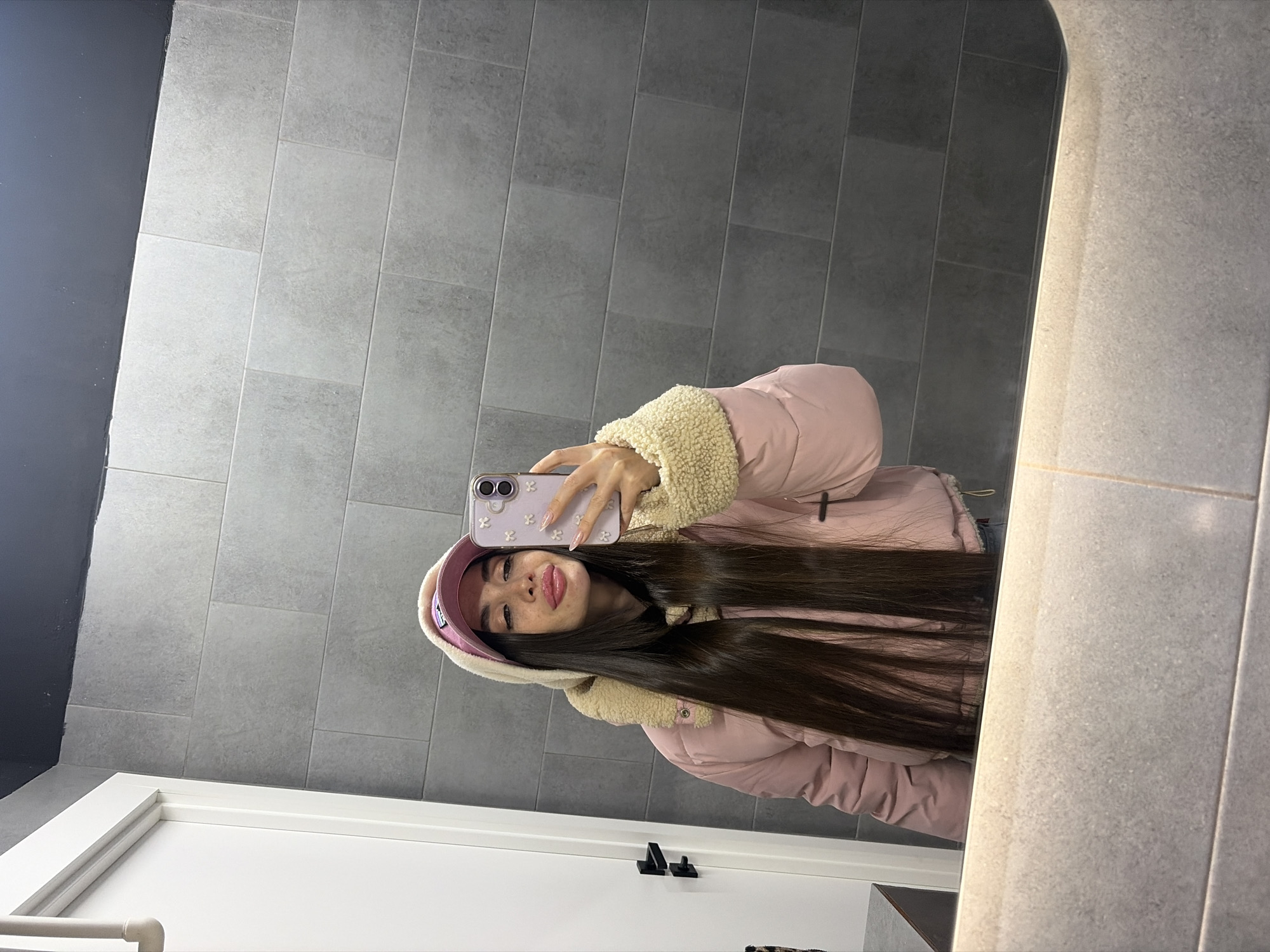 A young woman with long dark hair is taking a selfie in what appears to be a bathroom. She is wearing a pink puffer jacket with a cream-colored fleece collar and a matching pink baseball cap. She is holding a light purple smartphone decorated with white floral designs in her left hand. The background consists of gray rectangular tiles on the wall and a white door with a black handle. The location is unknown and cannot be deduced from the image.