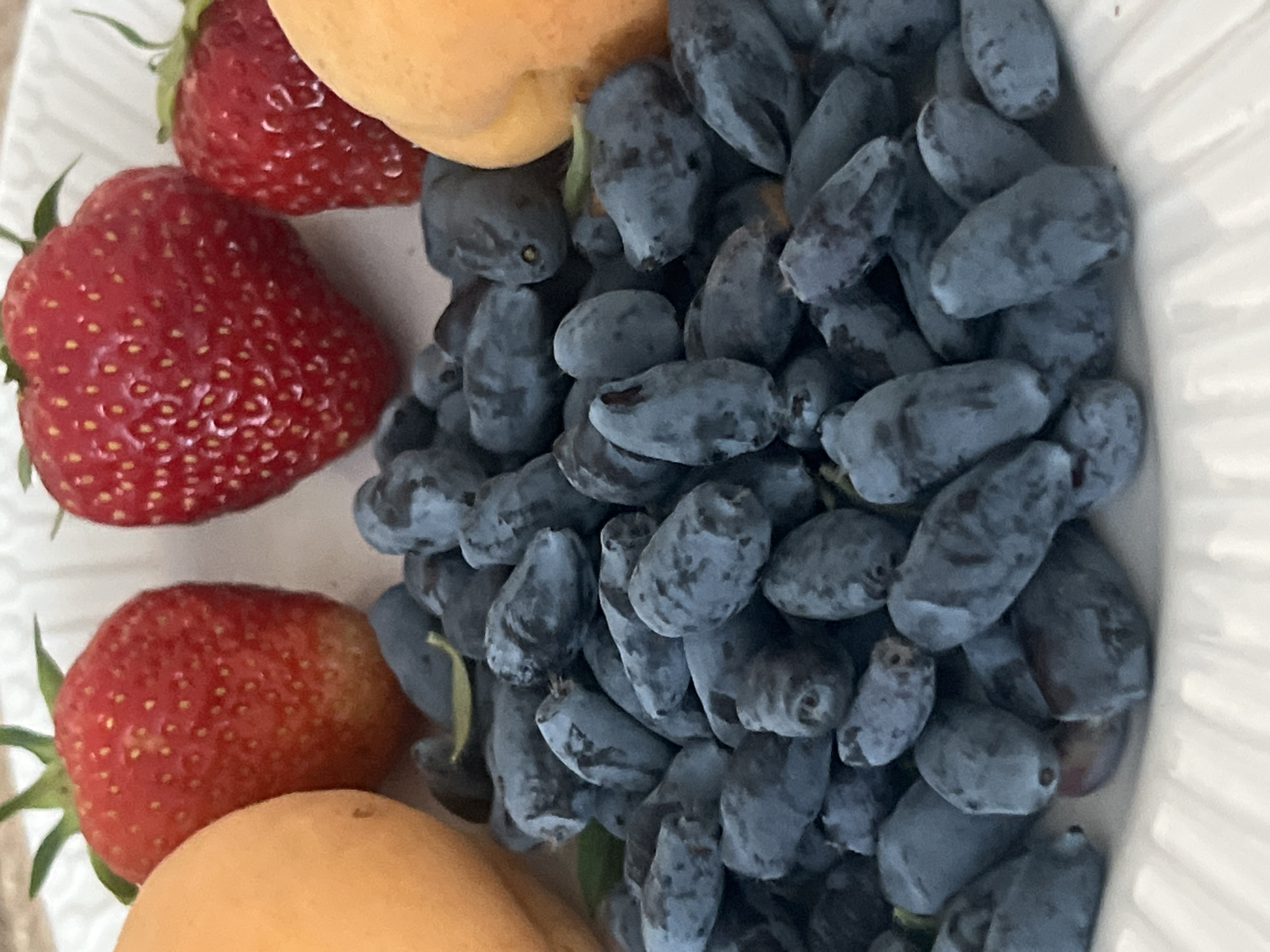 A close-up, overhead shot showcases a pile of dark blue, elongated berries, possibly honeysuckle, filling the foreground. To the left and right, the curved surfaces of two peaches or apricots are visible, one on the far left and another on the far right. In the background, three bright red strawberries are arranged with their green tops pointing upwards, one centered behind the berries and two slightly to the left and right. The fruits are presented on a white, textured surface, likely a plate. There are no people or discernible environmental features of Votkinsk, Russia, in this image.
