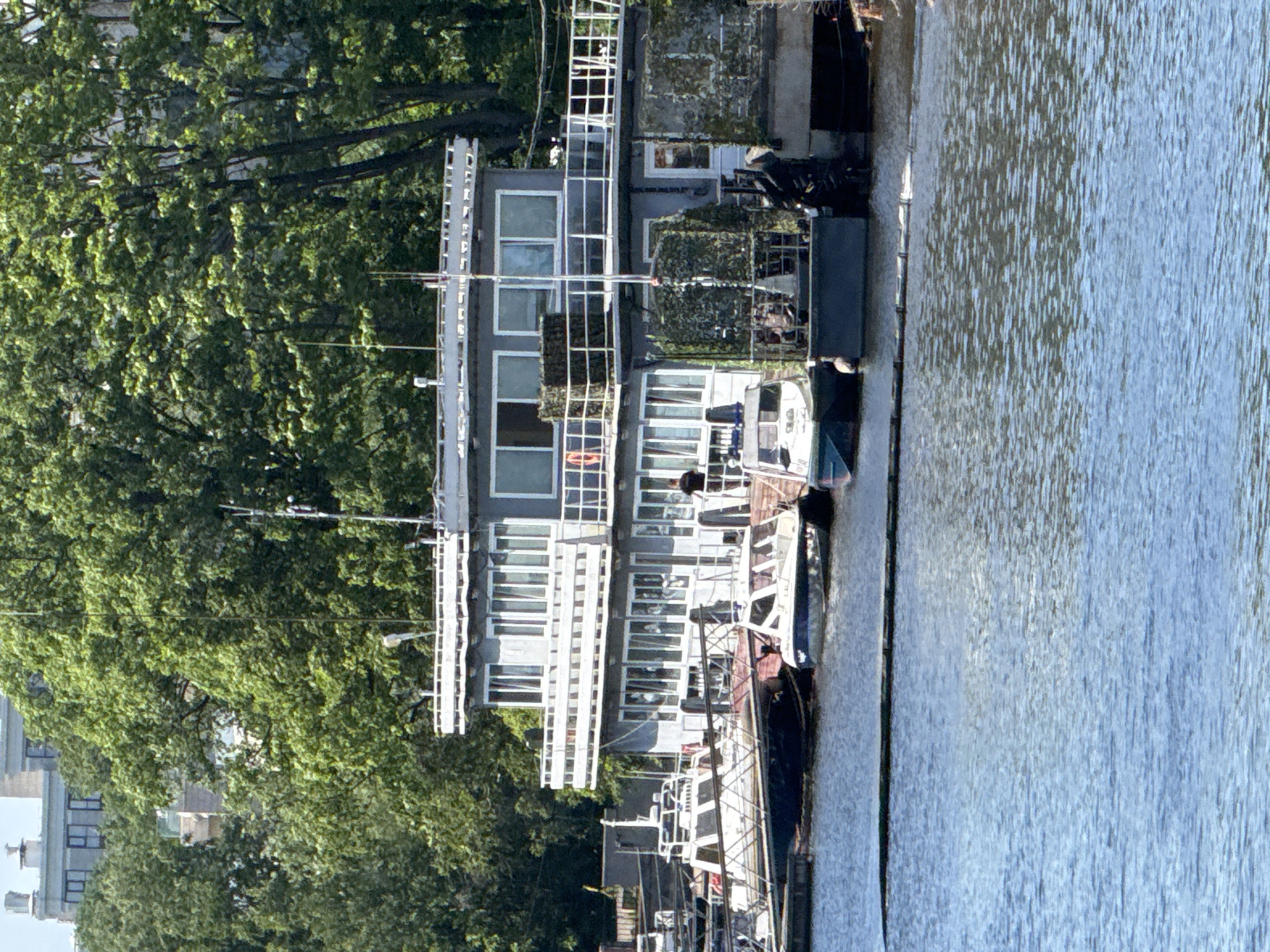 The image depicts a floating structure on a body of water, likely a river or canal in Saint Petersburg, Russia, given the architectural style of the distant buildings. The structure appears to be a two-story building with a gray exterior and numerous windows on both levels. The upper level features a balcony or deck with a railing, and several antennae are visible on the roof. Several boats are moored alongside the floating building, including a white boat with a dark hull and a smaller white boat with a dark blue hull. One person is visible on the deck of the floating structure, and another person stands on a dock. The environment is characterized by lush green trees in the background, suggesting a park or a densely wooded area adjacent to the waterfront. The water is calm, reflecting the bright sky.