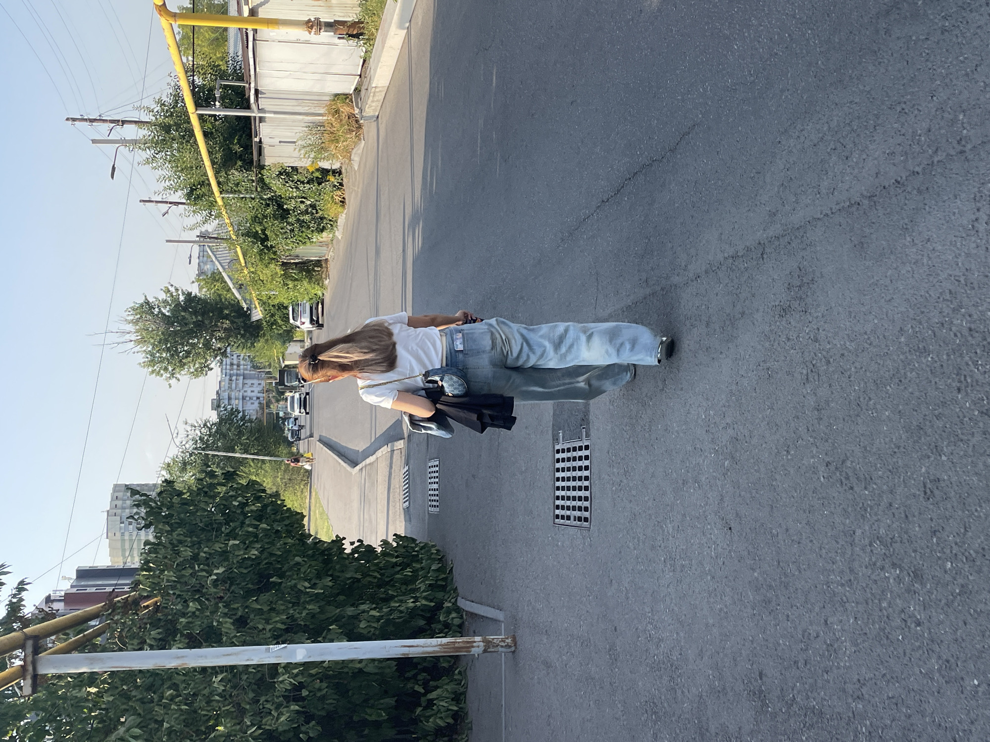 A woman with long, blonde-streaked hair, wearing a white t-shirt and light blue wide-leg jeans, walks away from the camera down an asphalt road in a city. She is carrying a dark jacket over her left arm and a small, rounded blue handbag over her right shoulder. The road is lined with green trees on the left and a corrugated metal fence on the right, with a yellow gas pipe running above the fence. In the background, several apartment buildings are visible, along with utility poles and wires. The scene appears to be in a residential or mixed-use area of Almaty, Kazakhstan, likely during the daytime with clear skies. There are two metal grates on the sidewalk to the left of the woman. Several cars are driving in the distance on the road.