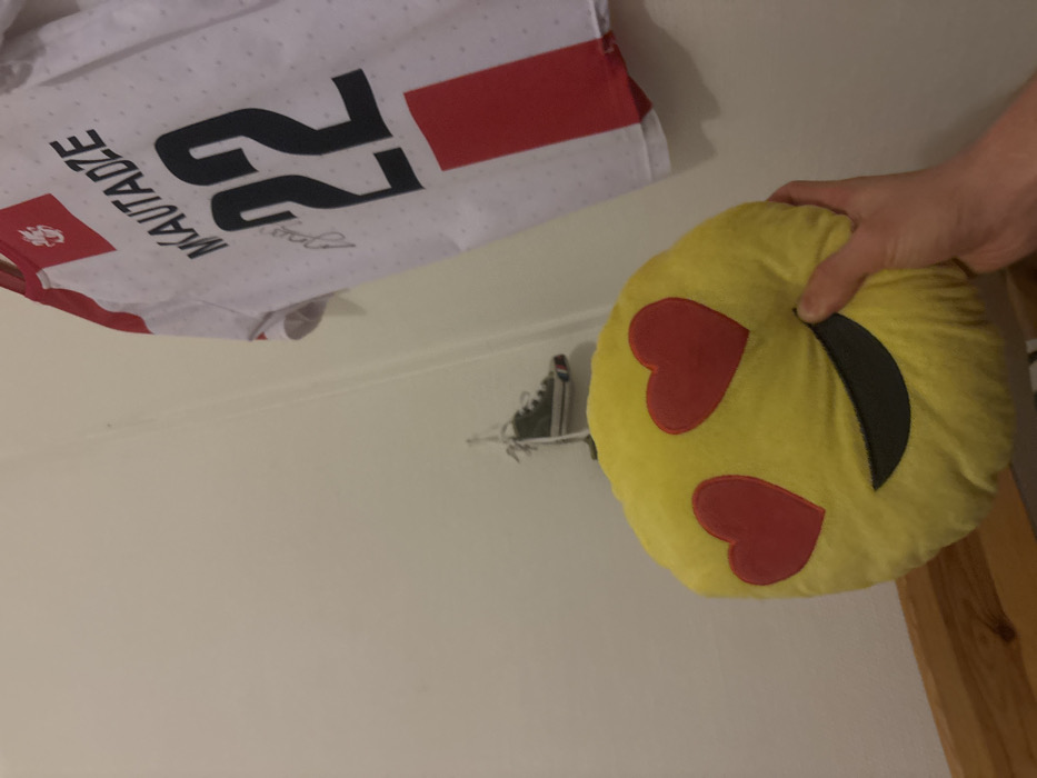 A hand holds a round, yellow emoji pillow with red heart-shaped eyes and a black smiling mouth. Above, a white sports jersey with the name "MAUTADZE" and the number "22" is partially visible. In the background, a white wall includes a small shelf displaying a gray sneaker. The edge of a wooden doorframe appears on the right.