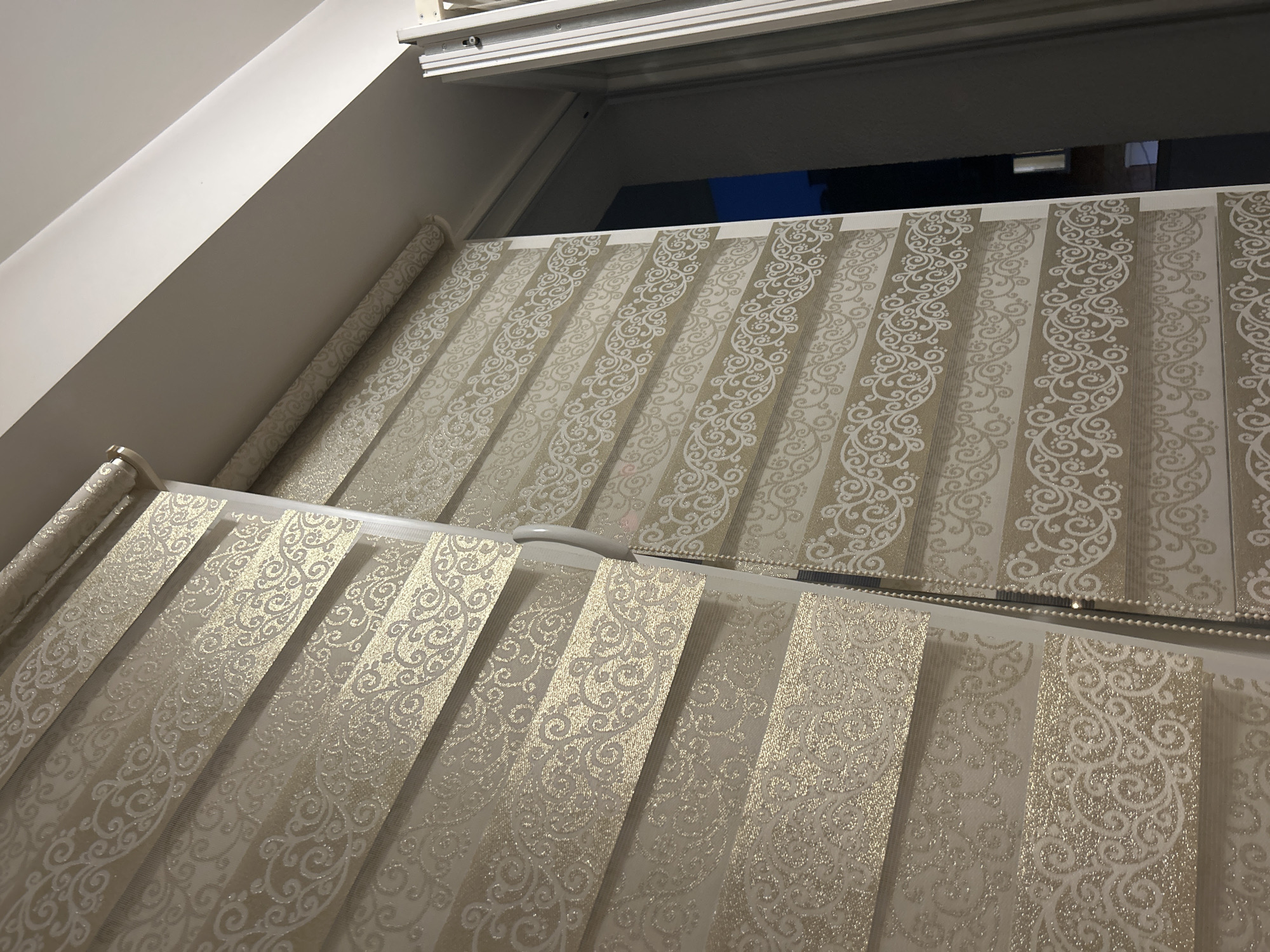 The image shows a close-up of two layered roller blinds with a decorative pattern. The blinds are positioned in front of a window. The left blind has a pattern of gold glitter and a swirling white design on alternating transparent and opaque stripes. The right blind has a similar design but the pattern is white on alternating transparent and opaque stripes. The blinds are partially rolled up. The environment appears to be indoors, possibly a residential or commercial space, given the style of the blinds and the presence of a window with a view of the outdoors, showing a dark sky and some building lights. The lighting in the room is artificial and appears to be from above, casting subtle shadows on the blinds. The location is unknown.