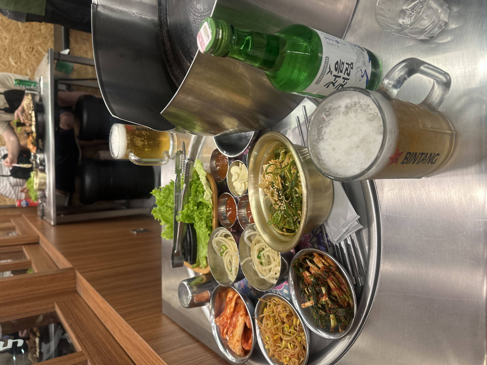 The image shows a table-top Korean BBQ setup. In the foreground, a mug of beer with a foamy head sits on the table. Next to it is a green bottle of Chum Churum soju. A metal tray holds various small dishes of Korean side dishes, including kimchi, bean sprouts, and what appears to be pickled greens. Lettuce leaves and tongs are also on the tray, likely for wrapping grilled meat. In the background, other diners are visible, and a metal grill is present on the table. The overall environment appears to be a restaurant.