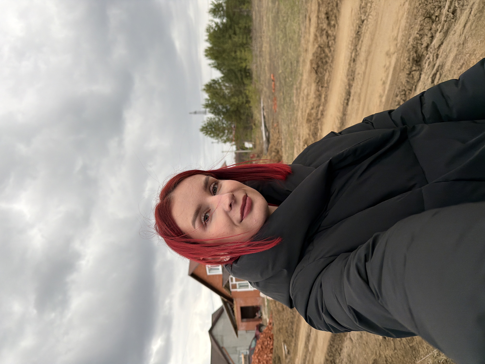 A woman with dyed red hair, wearing a black puffer jacket, is taking a selfie outdoors. In the background, there is a partially constructed house, a dirt road, and a cloudy sky. Trees can be seen in the distance. The scene appears to be located in Chaikovsky, Russia.