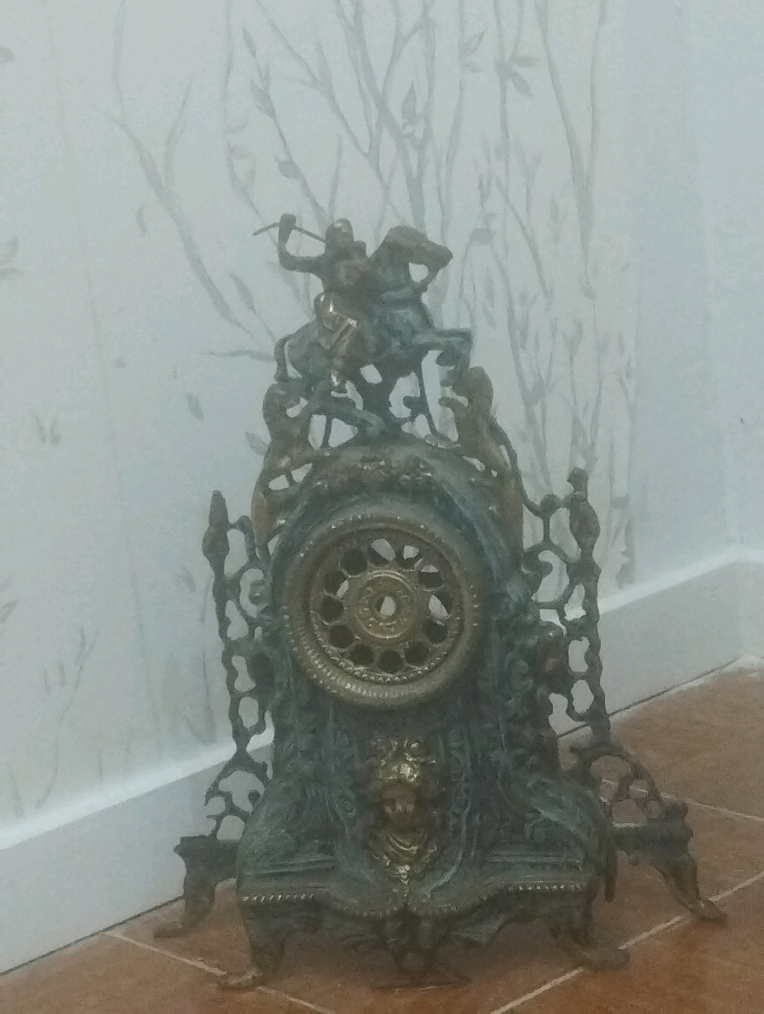 A metallic antique clock with ornate detailing is positioned on a tiled floor in front of a wall with a subtle, light-colored floral pattern. The clock features a central circular dial surrounded by intricate scrollwork and figures. At the very top of the clock, a detailed sculpture depicts a person on horseback wielding a weapon. The clock's base is also elaborately designed with decorative elements, including what appears to be a lion's head motif. The lighting in the scene is soft and diffused, casting faint shadows. The image appears to have been taken indoors. The location provided, Gondomar, Portugal, is noted.