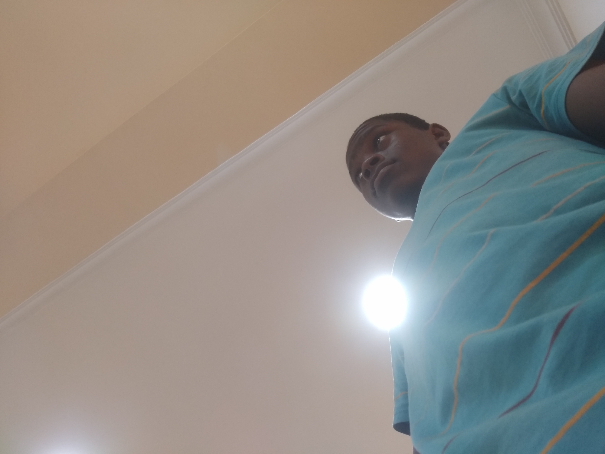 The image shows a person, presumably male, wearing a light blue t-shirt with thin yellow and maroon stripes. The person is positioned looking up and slightly to the left of the camera. The background consists of a plain, light beige ceiling. A bright light source is visible behind the person's shirt. The location is specified as Shangisha, Nigeria, but there are no elements in the image that definitively confirm this.